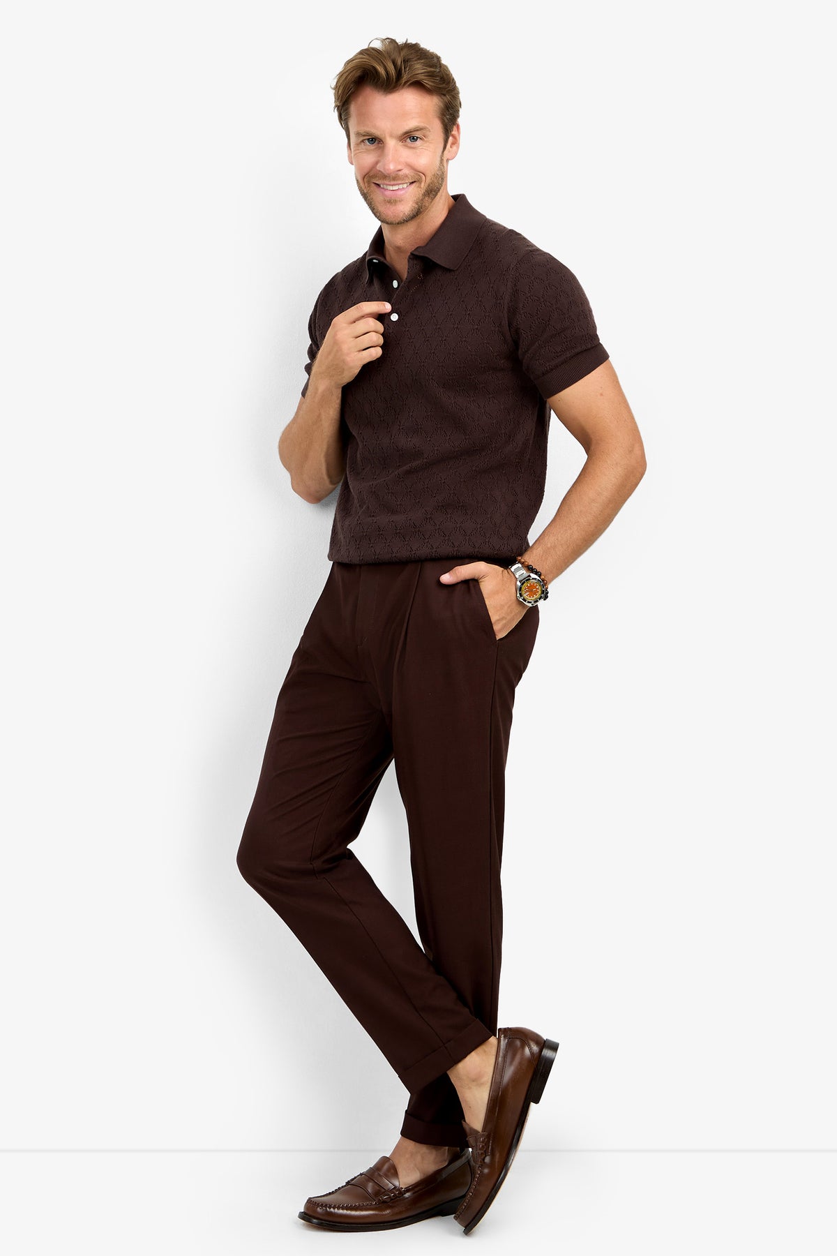 A man stands smiling, wearing the WAYLEN SS POLO in dark brown, paired with matching brown pants, brown loafers, and a watch. He has light brown hair and rests one hand in his pocket with a relaxed, confident pose.