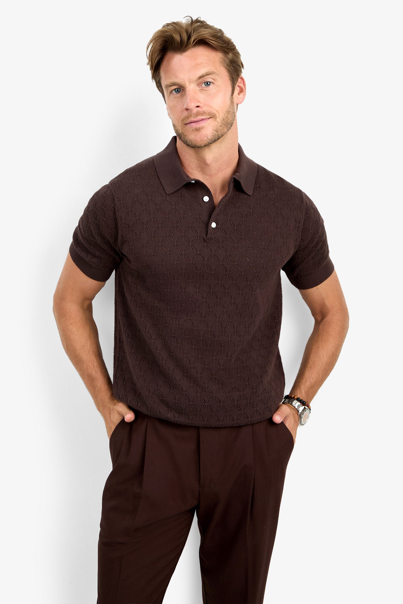 A man wearing the WAYLEN SS POLO in dark brown and matching brown trousers stands with his hands in his pockets against a plain white background.