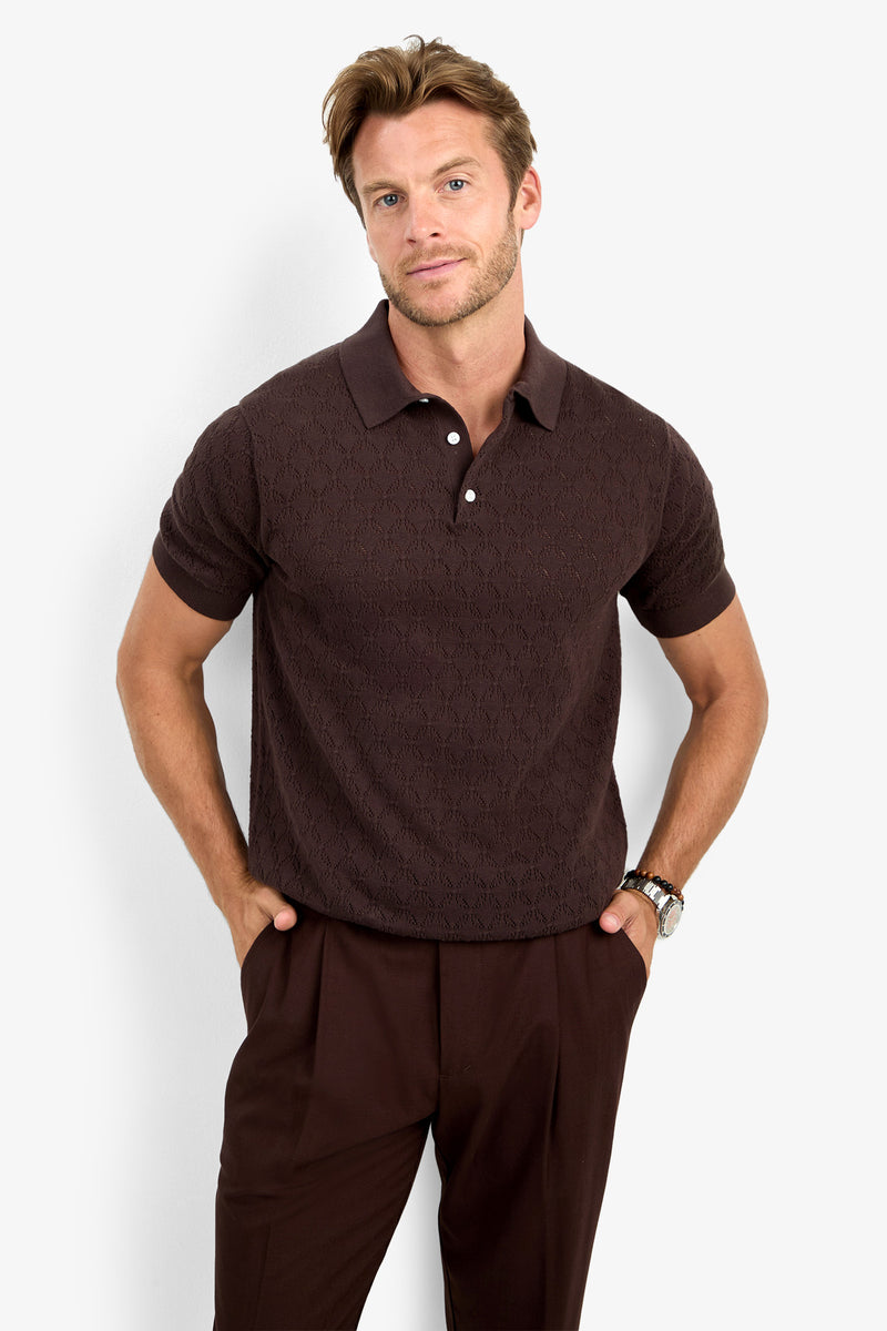 A man wearing the WAYLEN SS POLO in dark brown and matching brown trousers stands with his hands in his pockets against a plain white background.