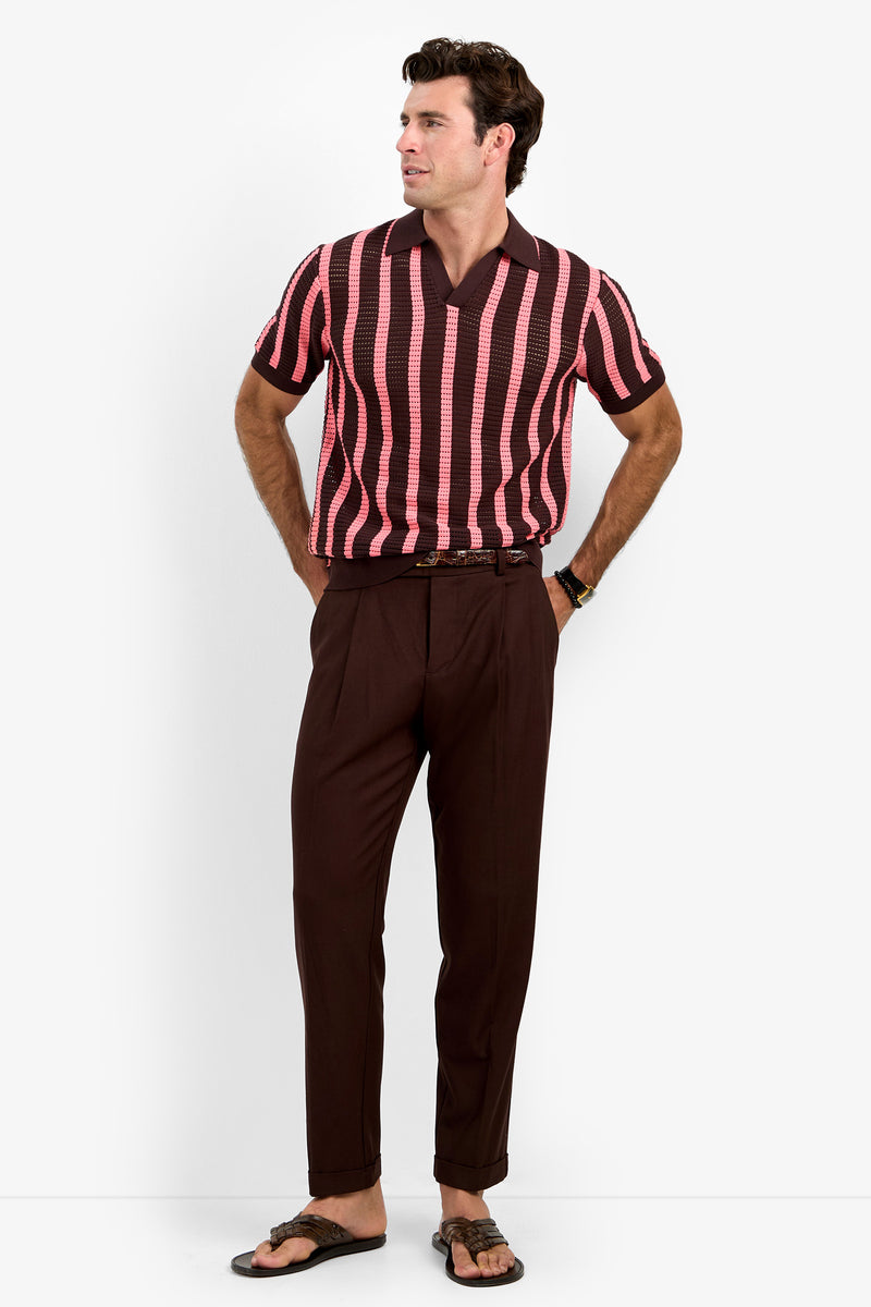 A man wearing the Alvin Mesh Polo—a short-sleeve, pink and brown striped knit shirt—paired with dark brown pleated trousers and sandals, stands with his hands in his pockets, looking to the side against a plain white background.