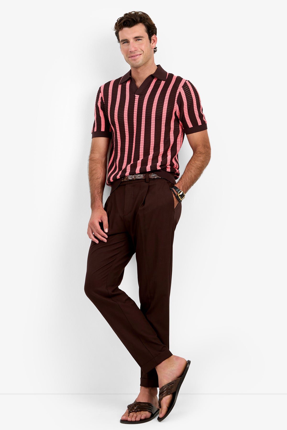 A man stands against a white background wearing the Alvin Mesh Polo, a pink and brown striped short-sleeve shirt. He pairs it with brown trousers, a belt, and brown sandals, smiling with his hands in his pockets.