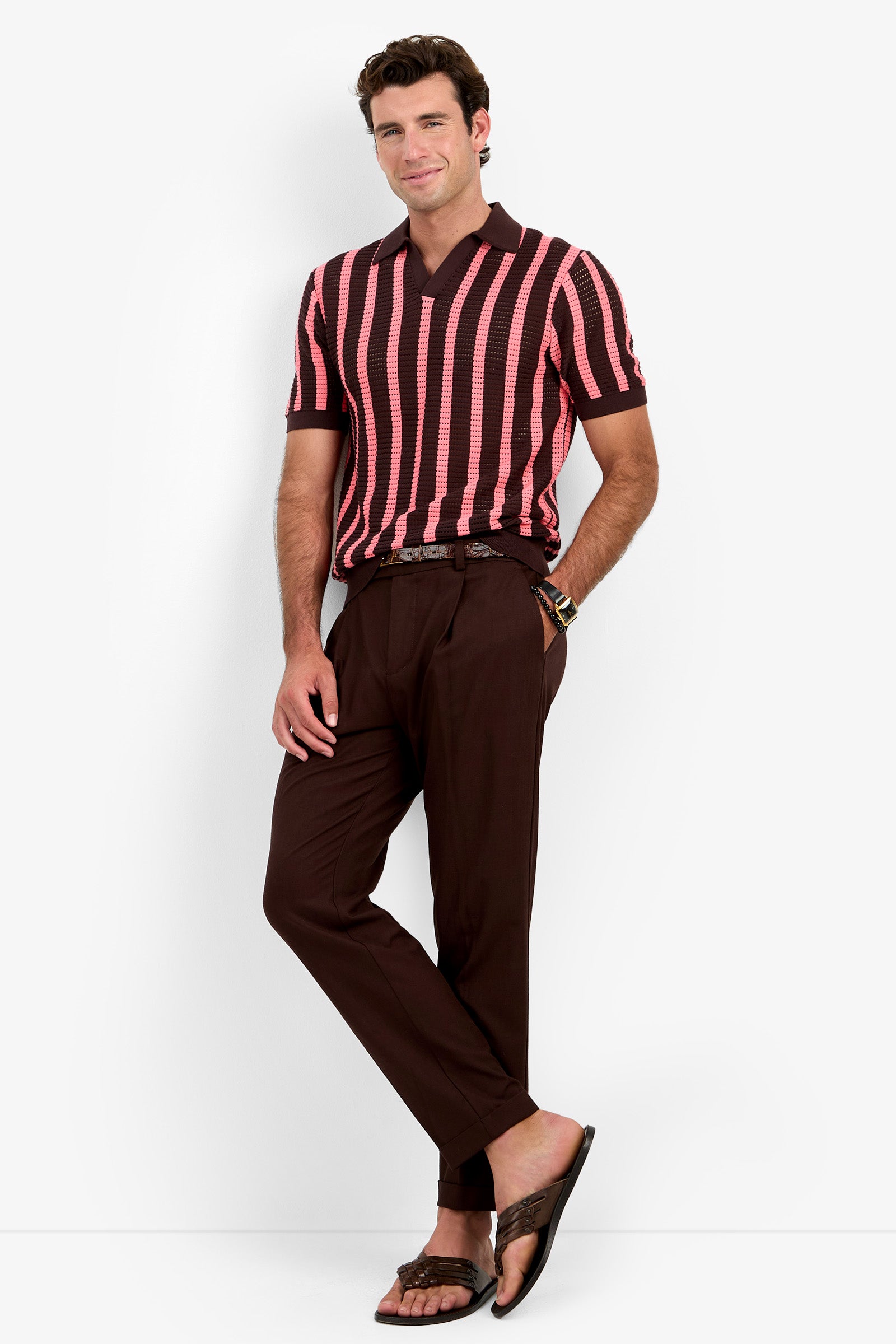 A man stands against a white background wearing the Alvin Mesh Polo, a pink and brown striped short-sleeve shirt. He pairs it with brown trousers, a belt, and brown sandals, smiling with his hands in his pockets.