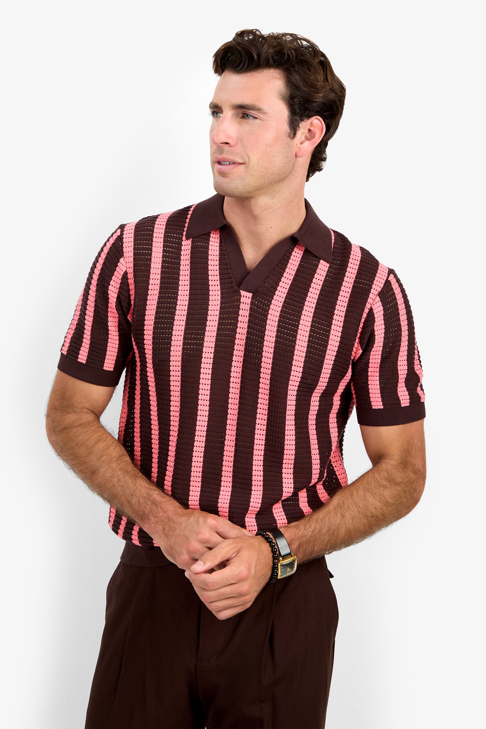 A man wearing the Alvin Mesh Polo, a short-sleeved knit shirt with brown and pink stripes and a collar, paired with dark brown pants, standing against a plain white background.