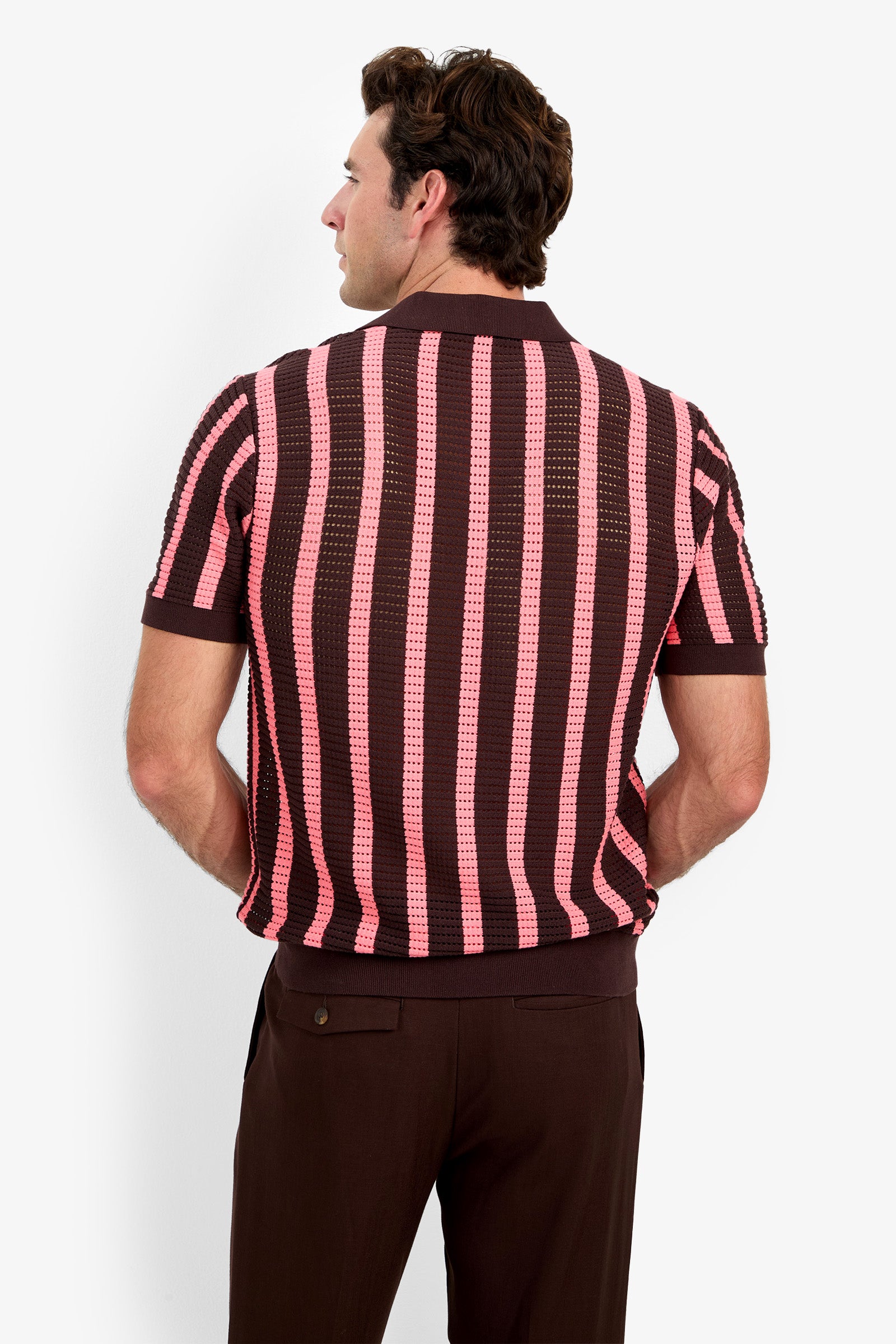 A man stands with his back to the camera, wearing the Alvin Mesh Polo—a short-sleeved, brown and pink vertically striped shirt—paired with dark brown trousers, set against a plain white background.