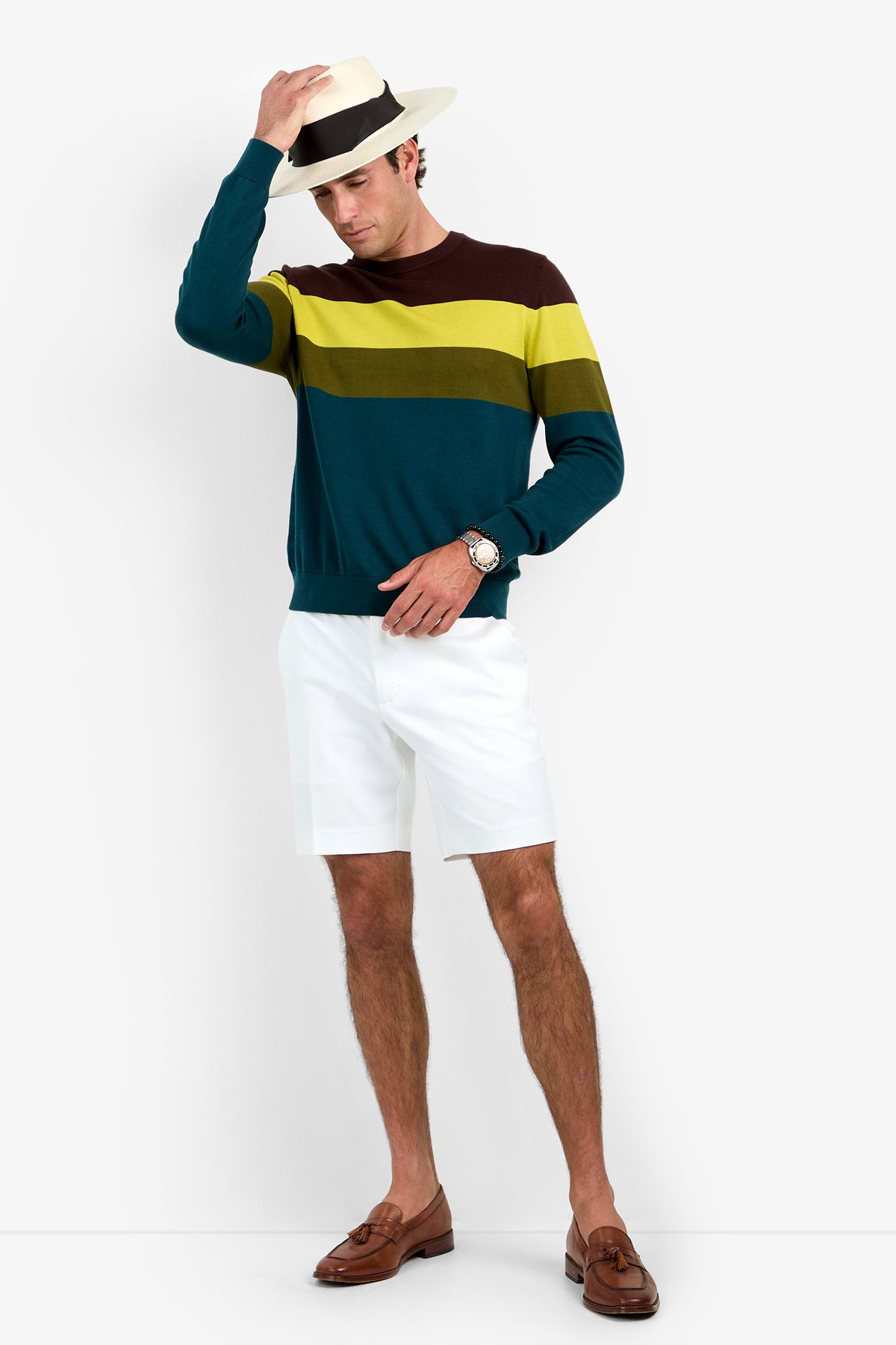 A man wearing the Roemer Crewneck sweater, white shorts, brown loafers, and a fedora hat touches the brim of his hat while standing against a plain white background.