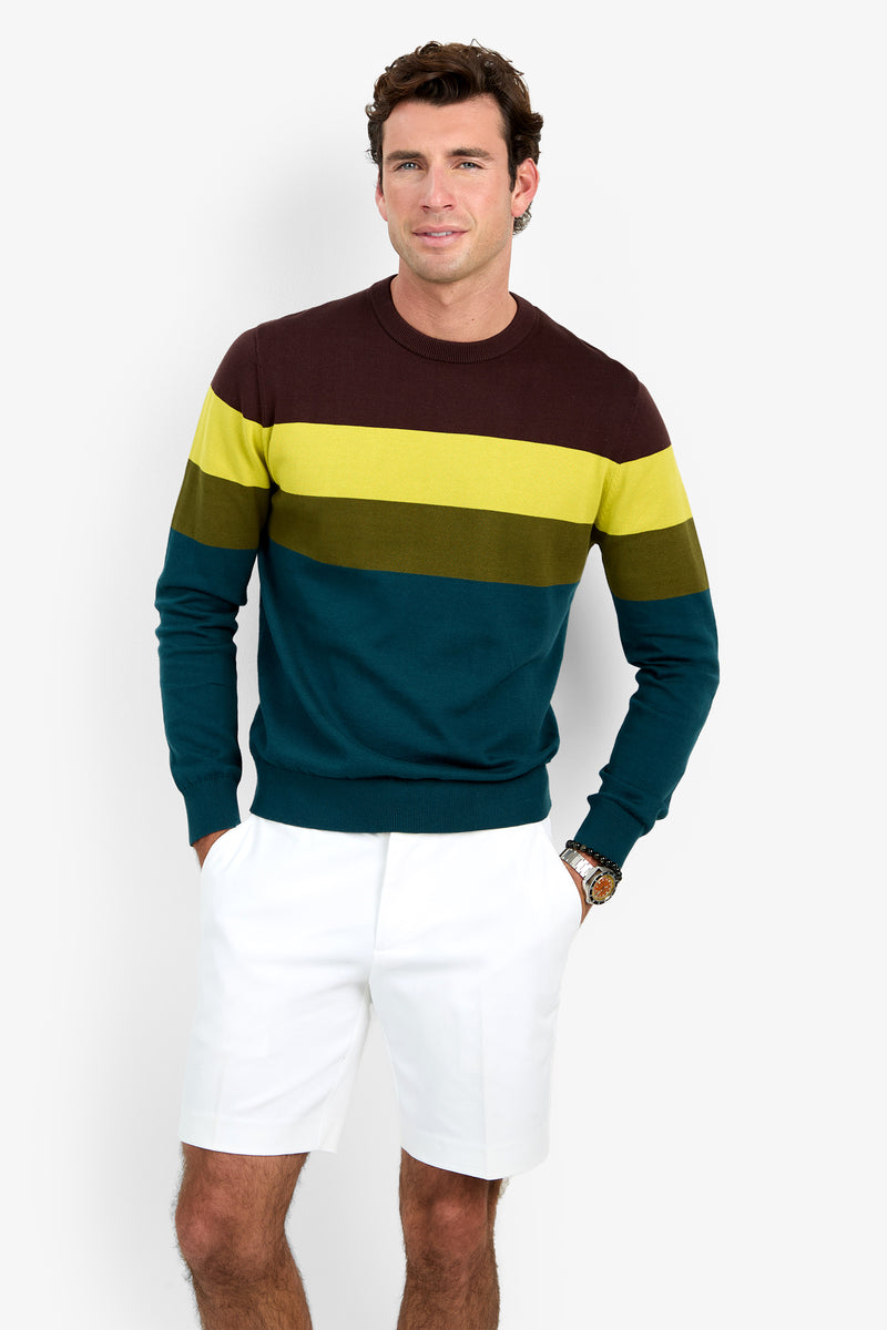 A man wearing the Roemer Crewneck, a striped sweater in shades of brown, yellow, and teal, pairs it with white shorts as he stands against a plain background with his hands in his pockets and smiles at the camera.