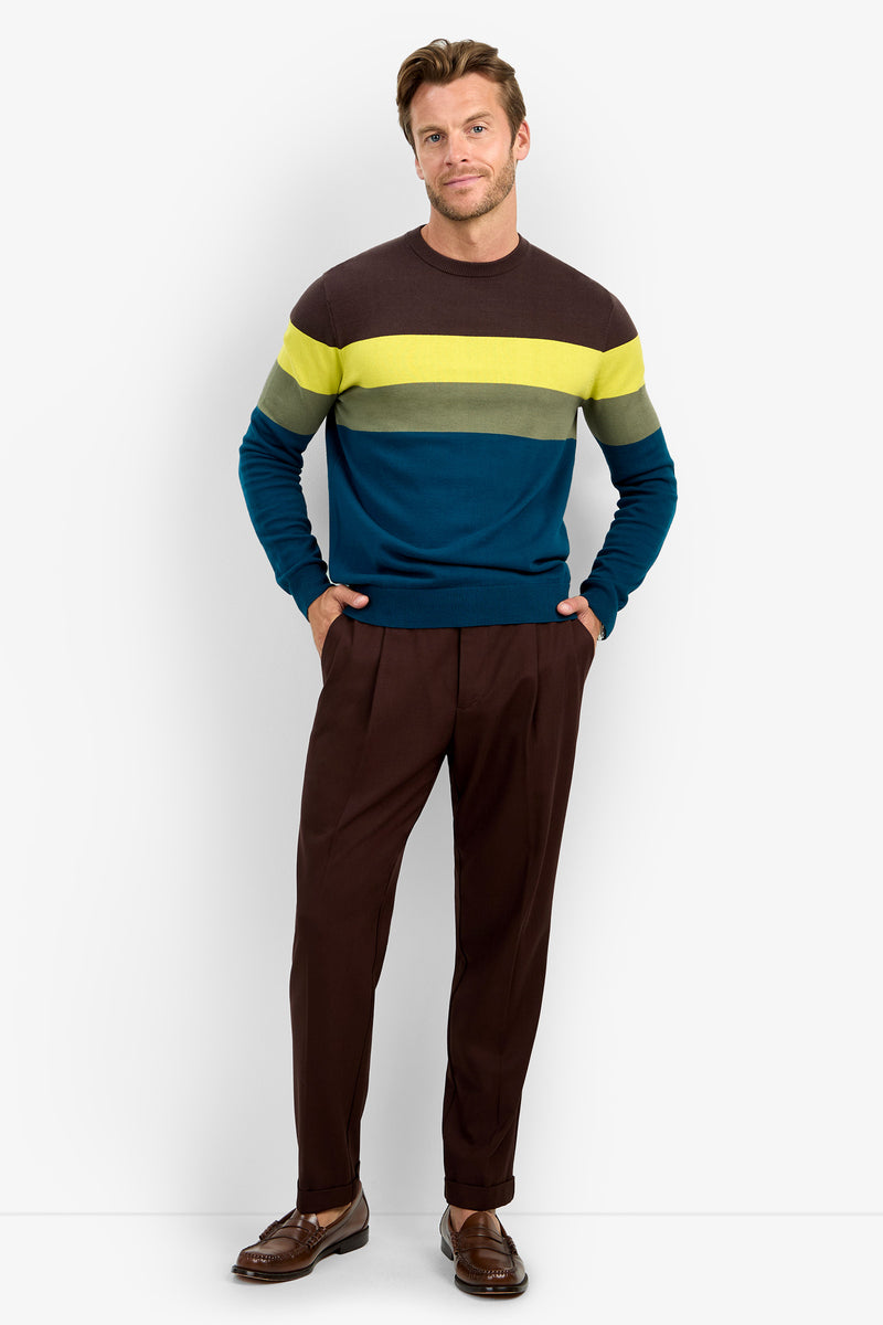 A man stands against a white background wearing the ROEMER striped sweater in brown, yellow, and blue tones, crafted from soft cotton fabric. He pairs it with brown pants and brown loafers, has his hands in his pockets, and smiles slightly.