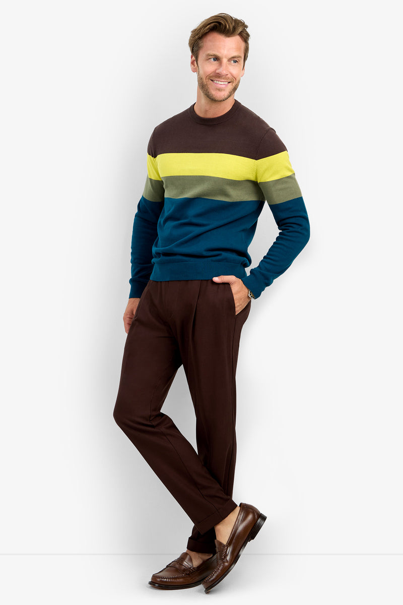 A man stands and smiles, wearing the ROEMER sweater—a color-block design crafted from 100% cotton with bold brown, yellow-green, and blue stripes. He pairs it with brown pants and dress shoes, posing casually with one hand in his pocket against a simple backdrop.