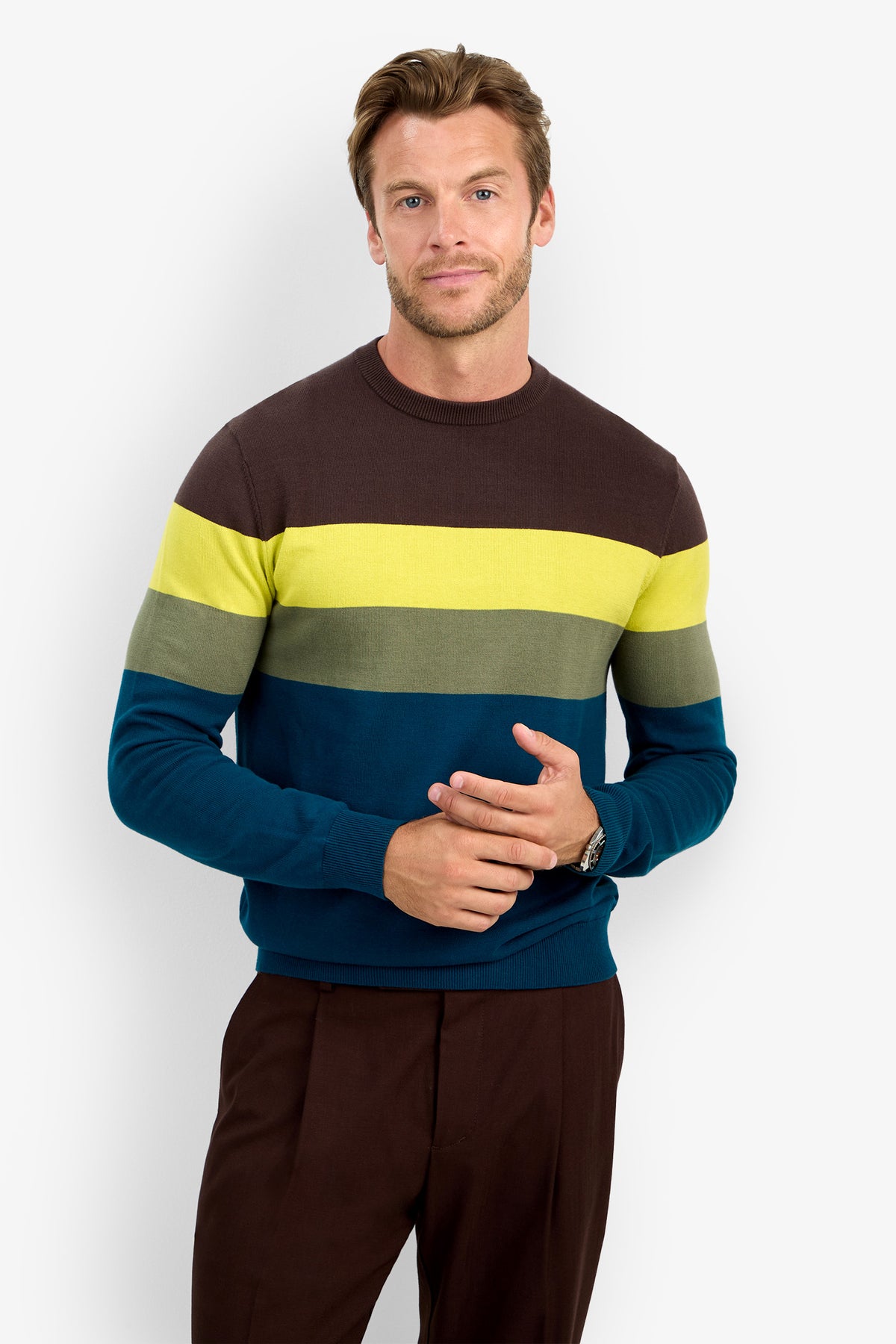 A man stands against a plain white background, with a slight smile and his hands loosely clasped, wearing the ROEMER 100% cotton sweater in brown, yellow, green, and blue stripes paired with brown pants.