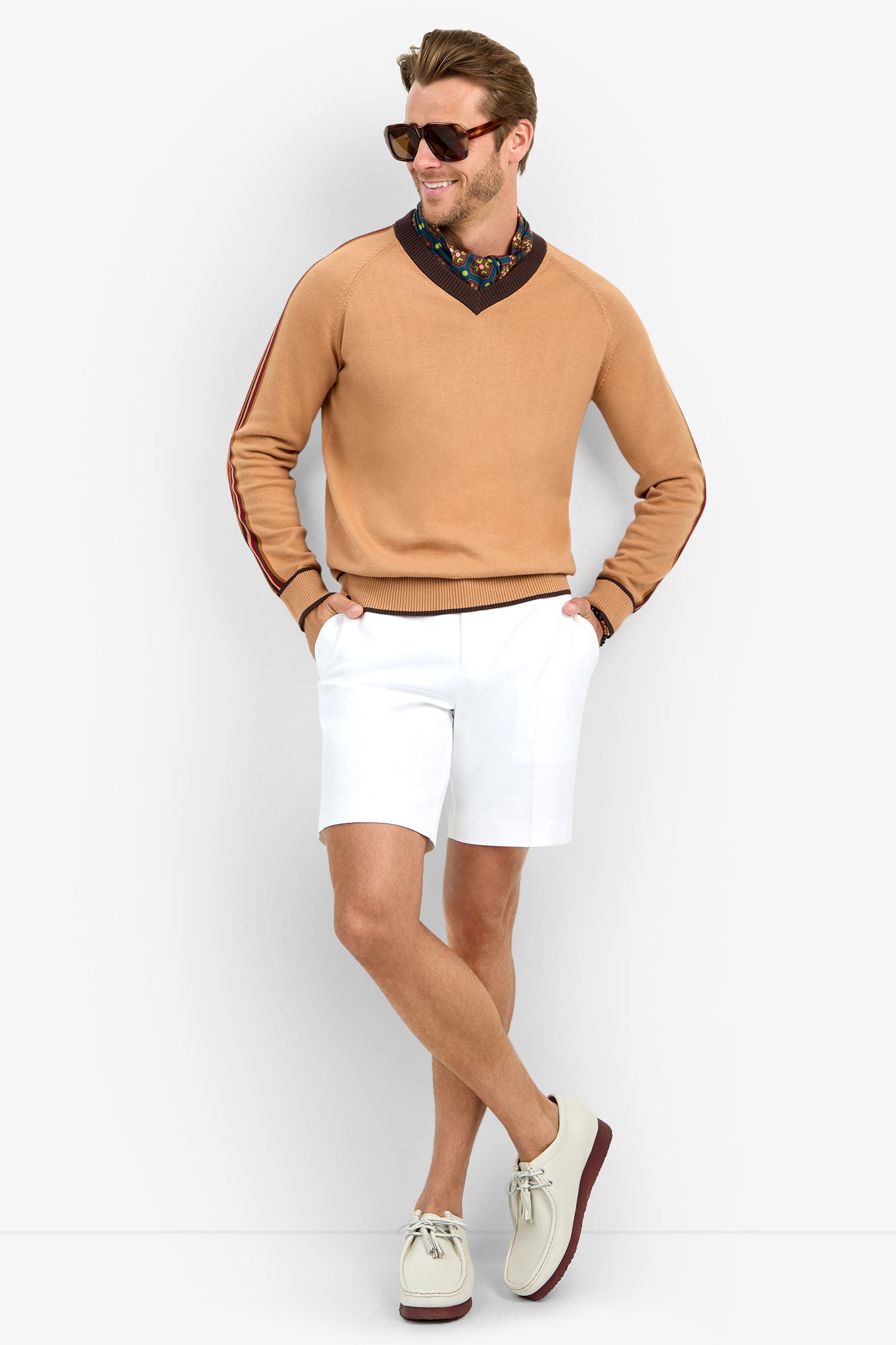 A man wearing HARVEY sunglasses, a tan sweater over a floral shirt, white shorts, and cream shoes stands smiling with his legs crossed against a plain background.