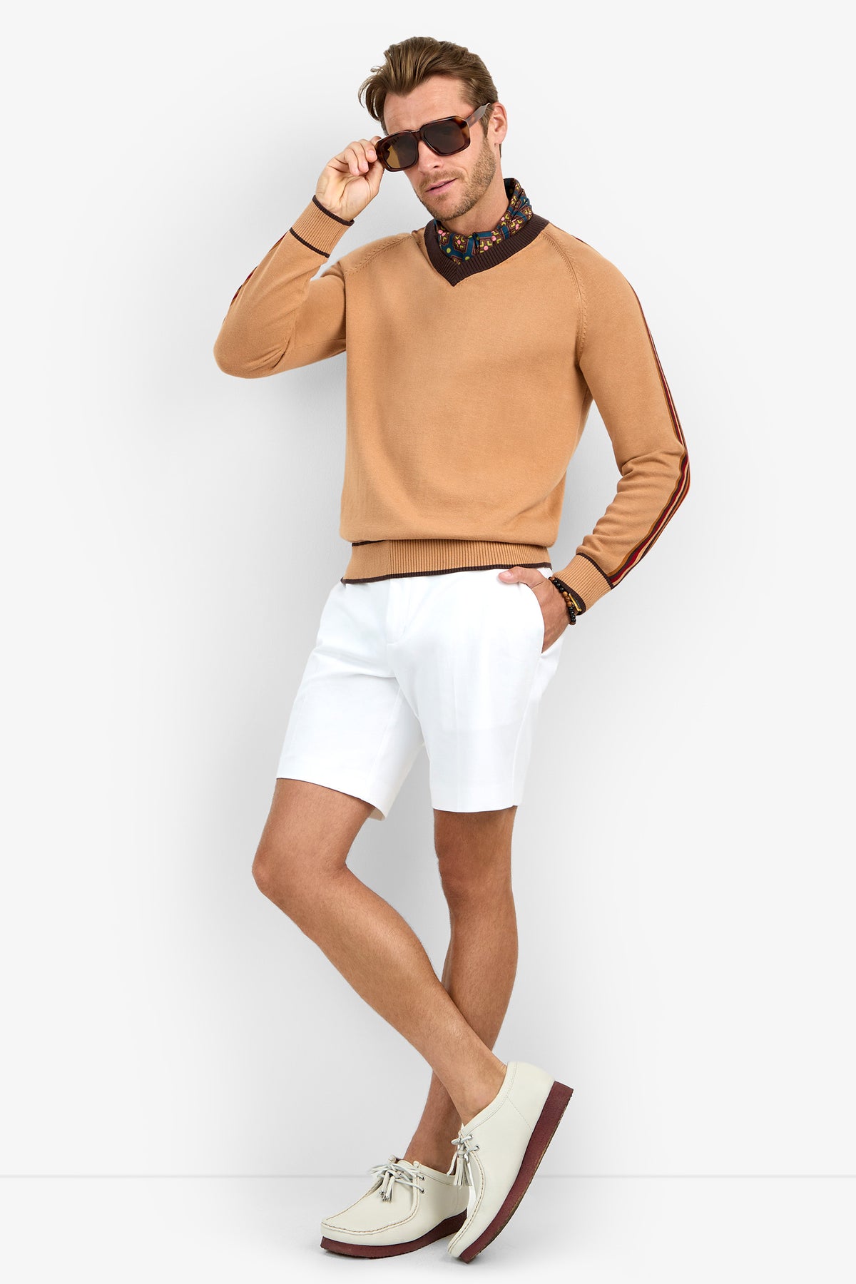 A man poses against a plain white background wearing the HARVEY sunglasses, a tan sweater with striped sleeves, a floral neck scarf, white shorts, and cream shoes.