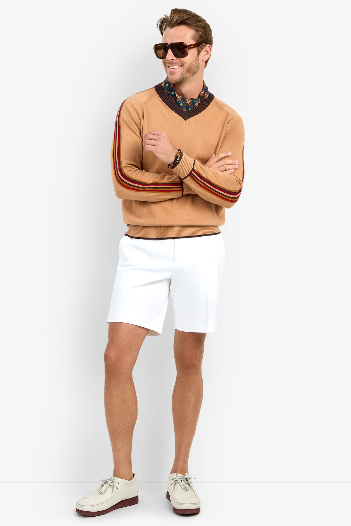 Wearing the HARVEY sweater with striped sleeves, a man in sunglasses pairs it with a floral collared shirt, white shorts, and cream shoes featuring brown soles. He stands smiling with his arms crossed against a plain white background.