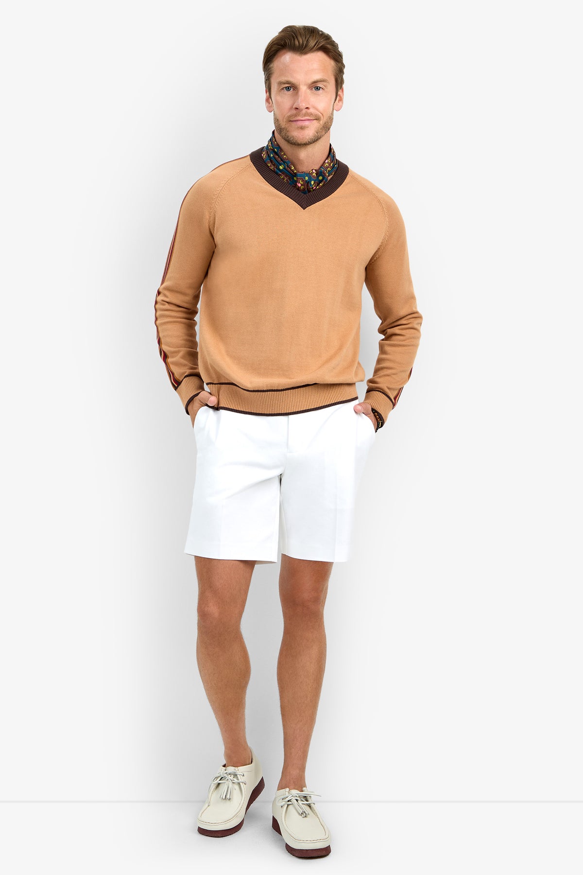 A man stands against a white background, hands in his pockets and smiling at the camera, wearing the HARVEY sweater in tan, layered over a floral-patterned shirt, paired with white shorts and cream shoes with brown soles.