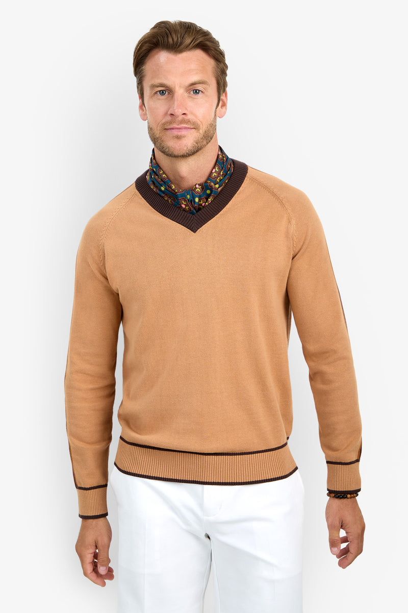 A man wearing the HARVEY tan sweater with dark brown trim, paired with a floral scarf and white pants, stands against a plain light background, facing forward with a neutral expression.