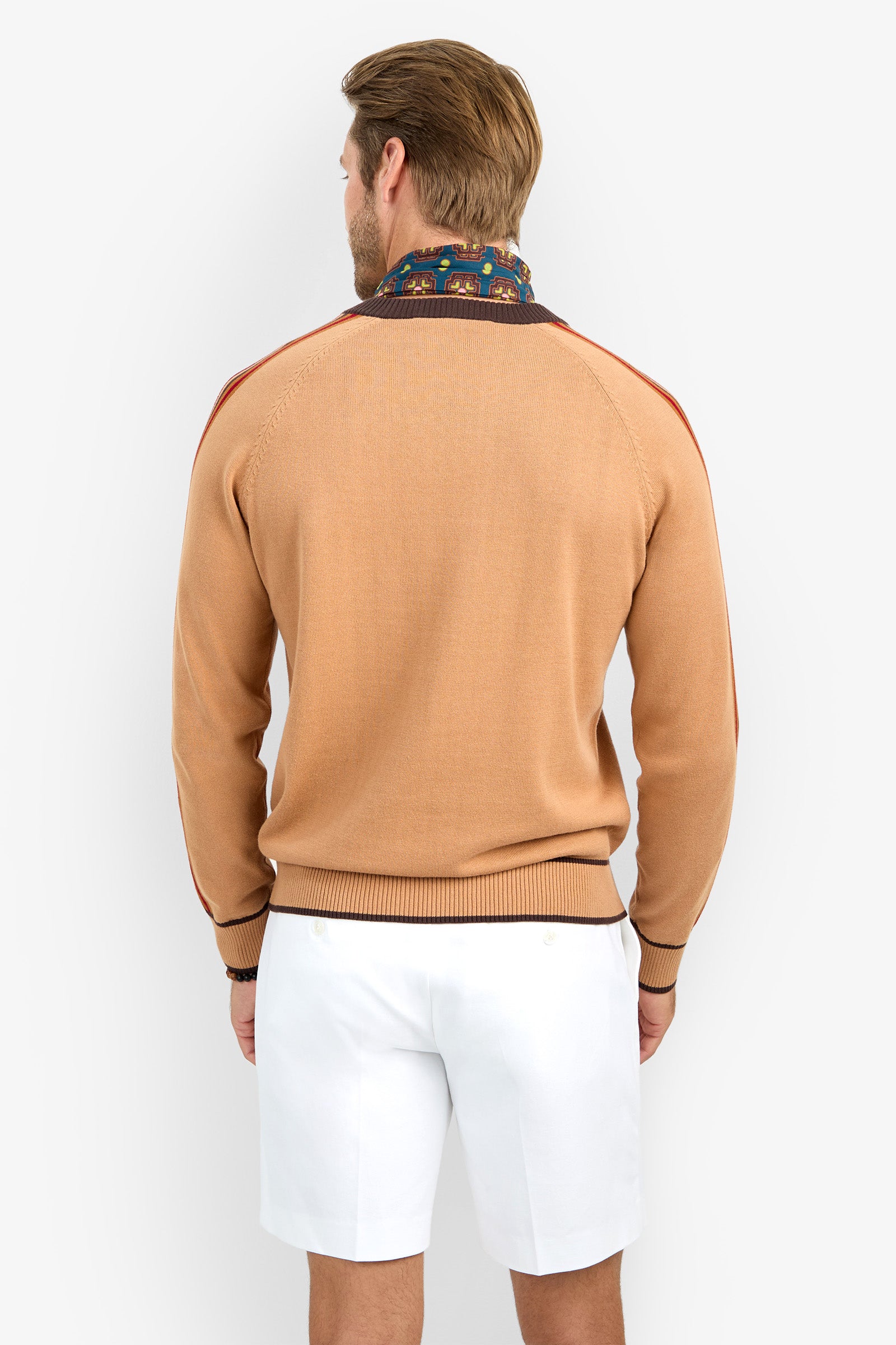 A man is standing with his back to the camera, wearing the HARVEY tan sweater with dark trim, a patterned scarf, and white shorts against a plain white background.