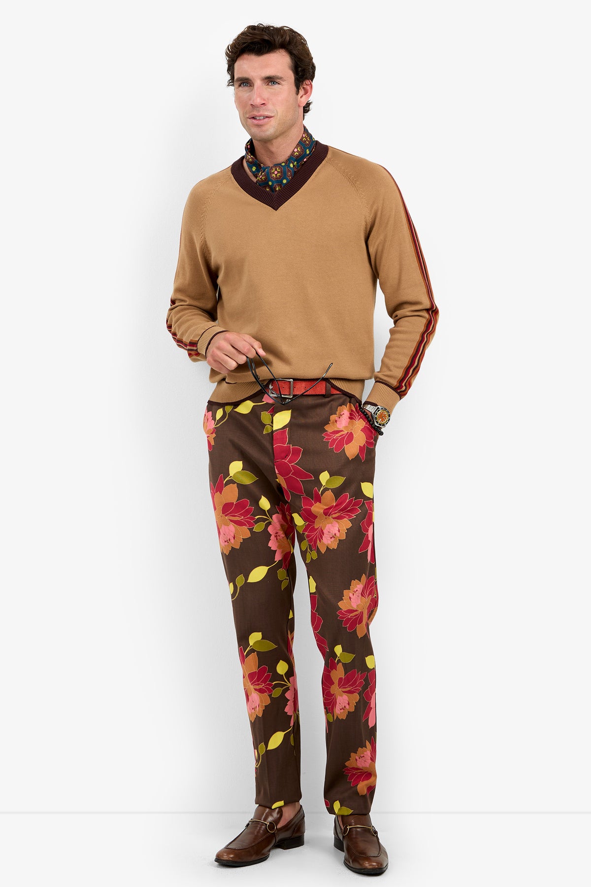 A man stands wearing the Harvey Sweater in tan over a floral shirt, paired with brown pants featuring large red and yellow flowers and brown loafers. He holds a belt and smiles.