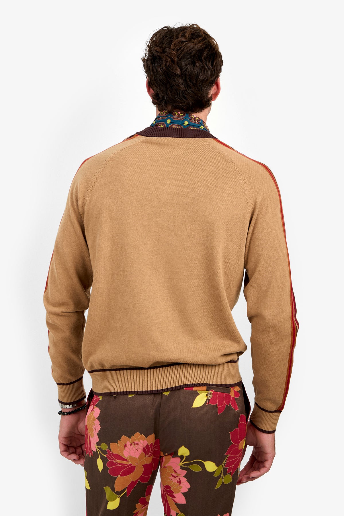 A person stands facing away against a plain white background, wearing the Harvey Sweater—a tan knit with red stripes on the sleeves and a patterned collar—paired with brown pants featuring colorful floral prints.