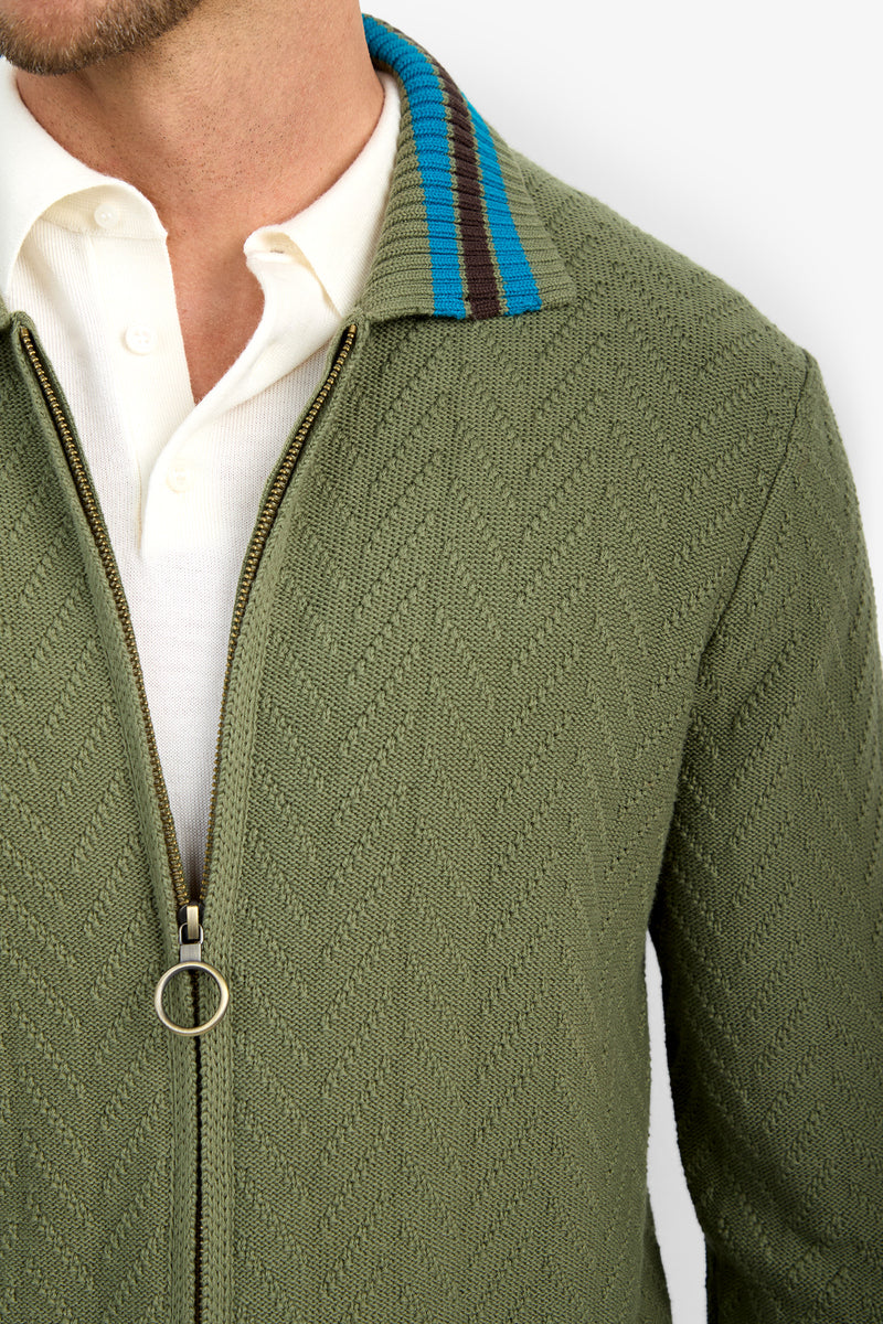 A close-up of a person wearing the ROBERTO ZIP CARDIGAN, an olive green textured zip-up jacket with a blue and brown striped collar, layered over a white shirt. Only the lower face, neck, and upper torso are visible.