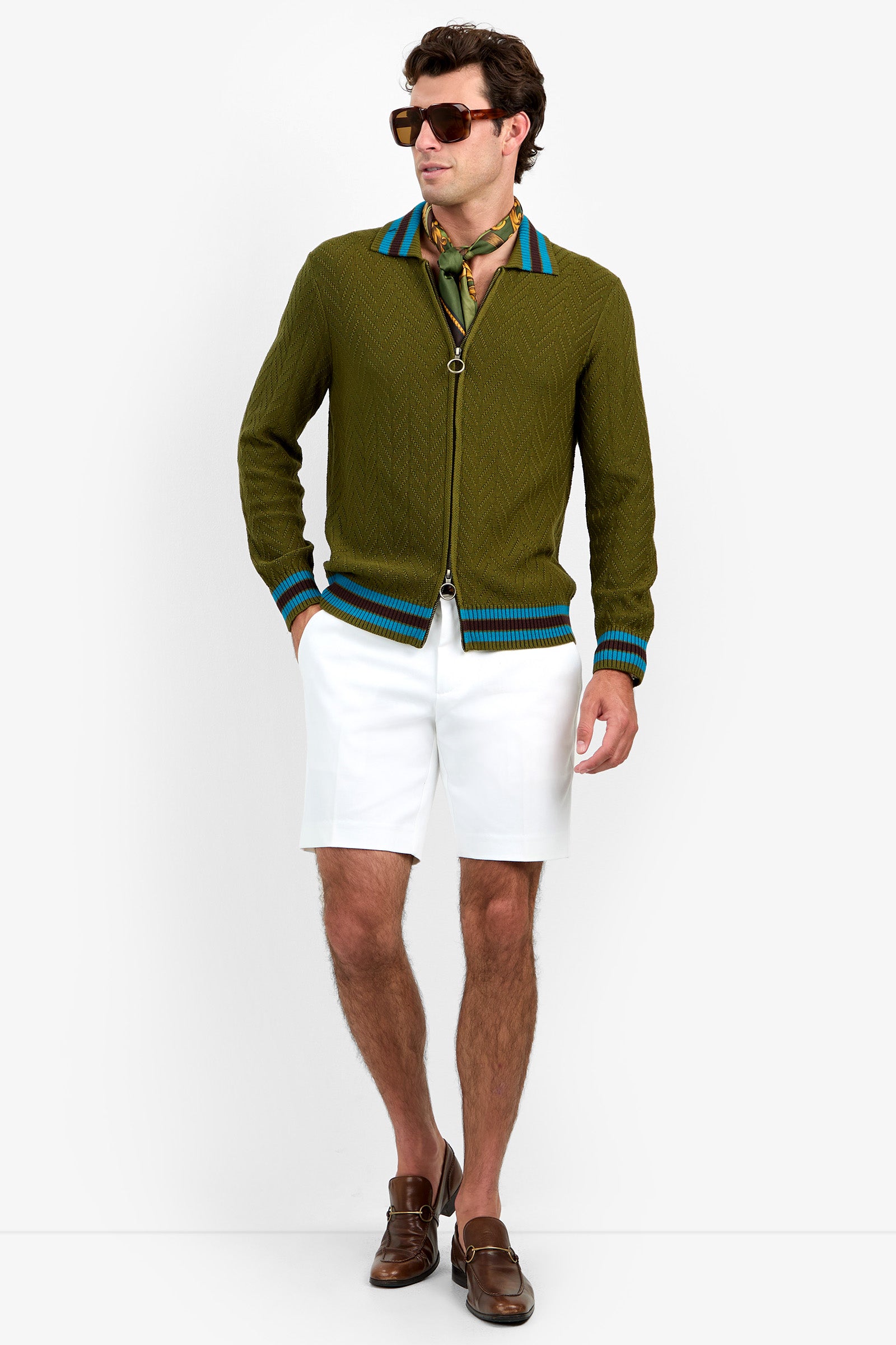 Wearing sunglasses and the Roberto Zip Cardigan—a green zip-up sweater with blue stripes—paired with a patterned neck scarf, white shorts, and brown loafers, a man stands against a plain white background.