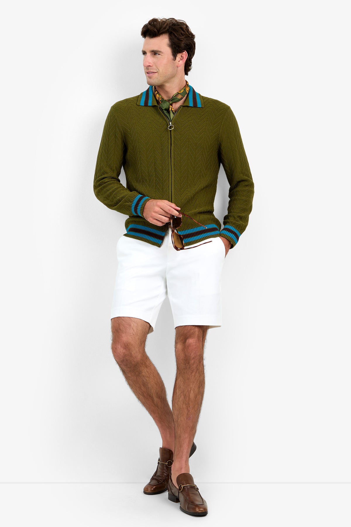 A man stands against a white background wearing the Roberto Zip Cardigan, which is green with blue stripes. He has paired it with a yellow scarf, white shorts, and brown loafers. Looking to the side, he poses with one hand in his pocket.