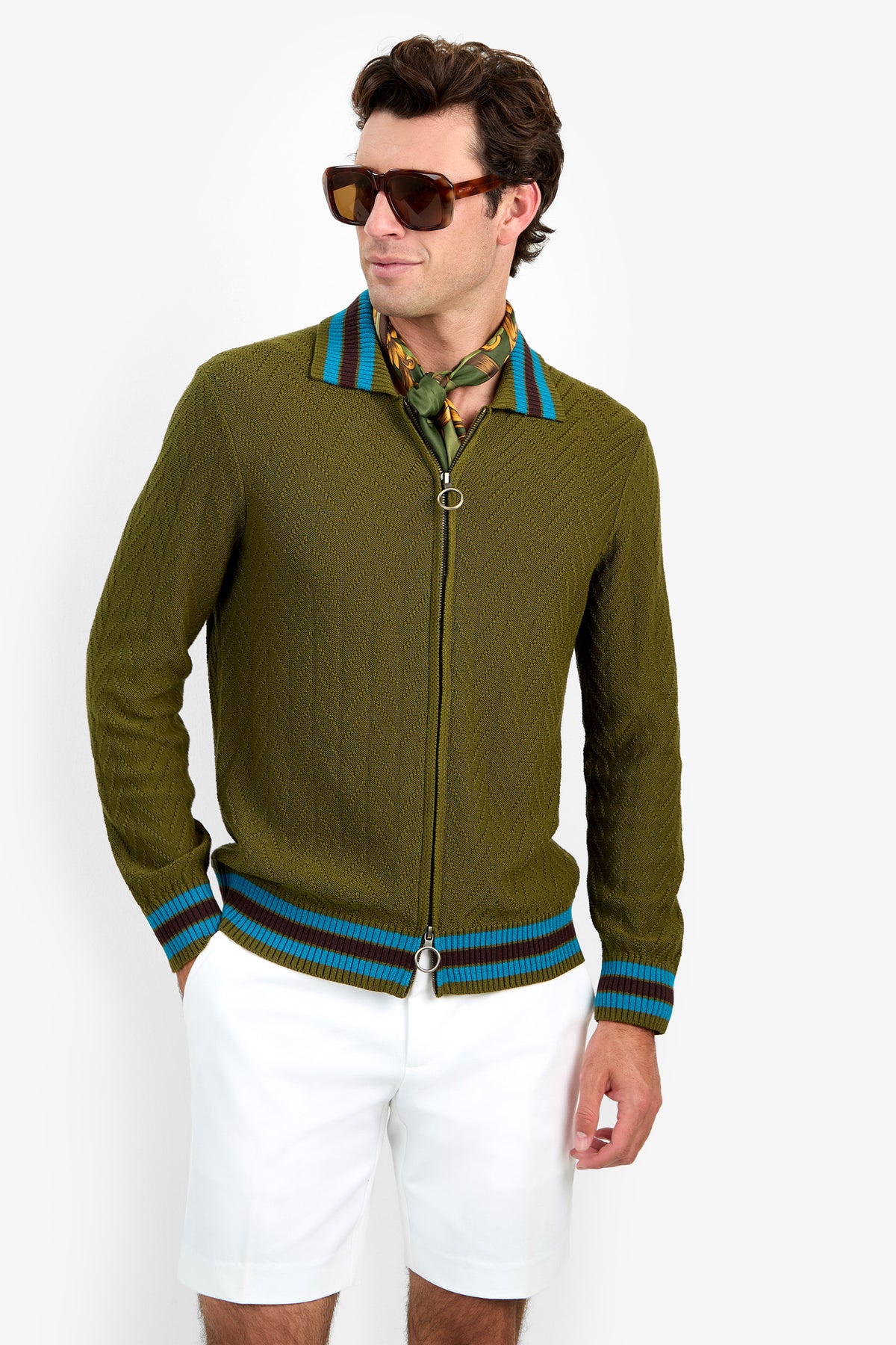 A man in sunglasses wears the Roberto Zip Cardigan, an olive green zip-up sweater featuring blue stripes on the collar and cuffs. He pairs it with a patterned scarf and white shorts while standing against a plain white background.