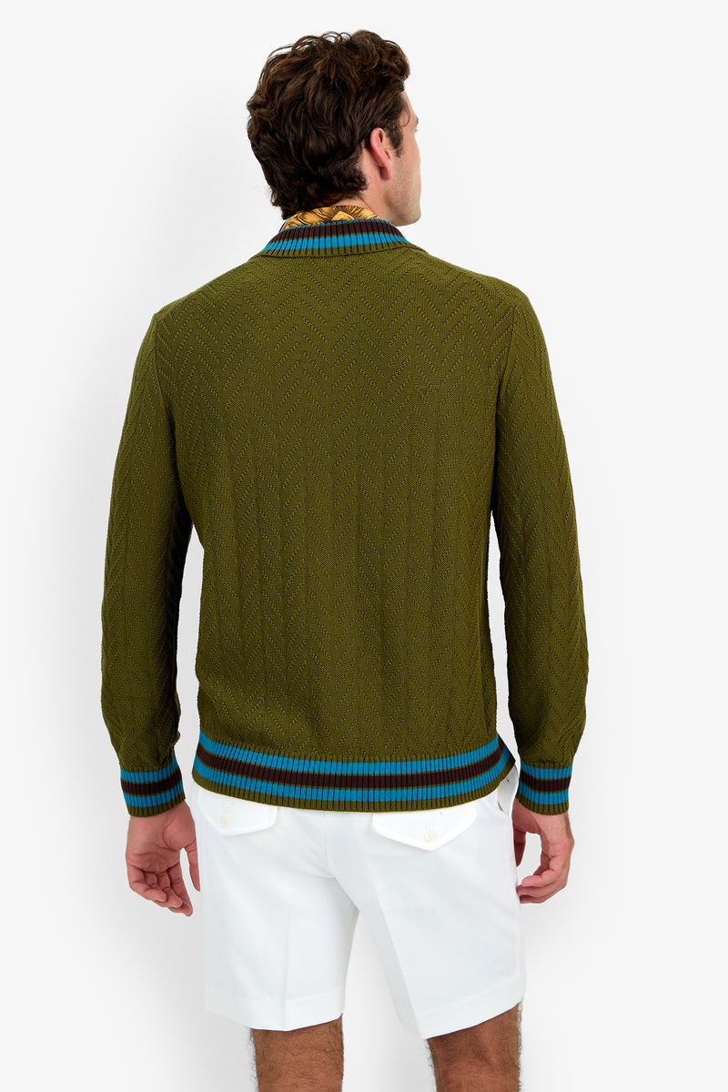 A man faces away, wearing the Roberto Zip Cardigan—an olive green sweater with blue and brown trim—and white shorts with buttoned back pockets, against a plain white background.