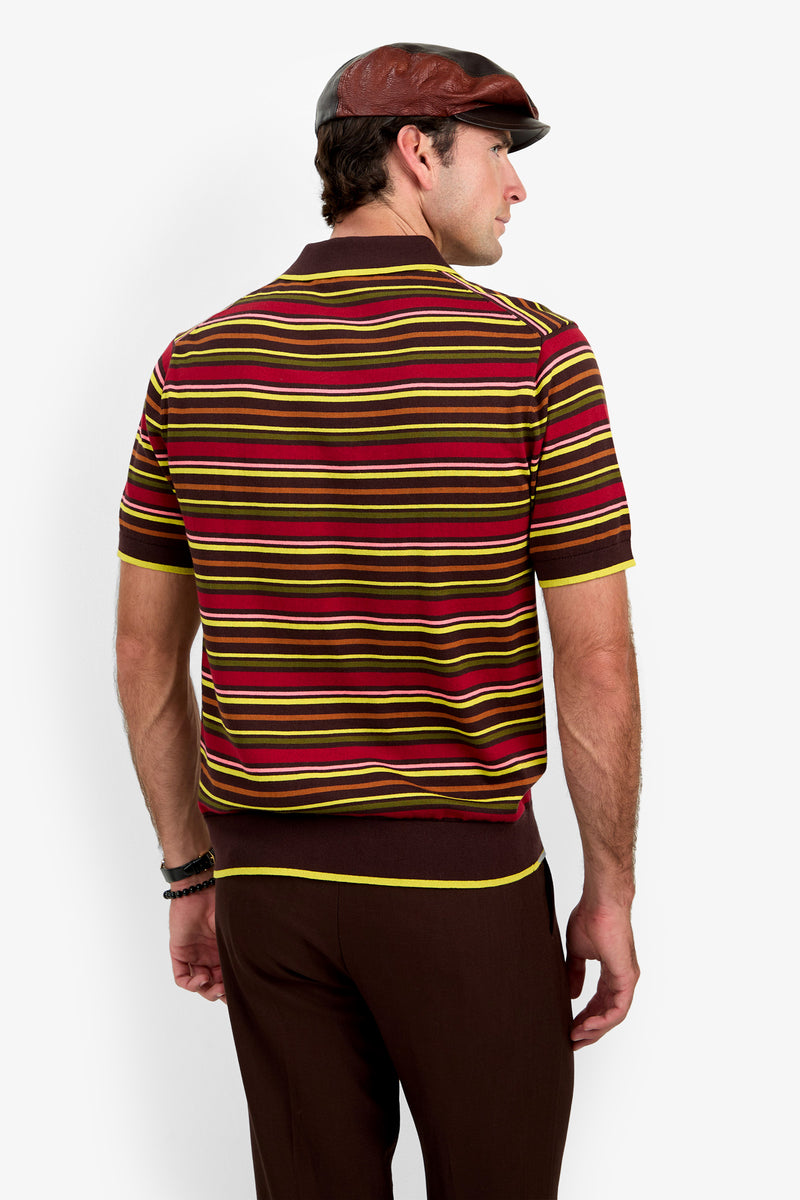 A man seen from behind wears the Bayard Polo—a red, yellow, and brown striped short-sleeve shirt—paired with brown pants and a brown flat cap, standing against a plain white background.