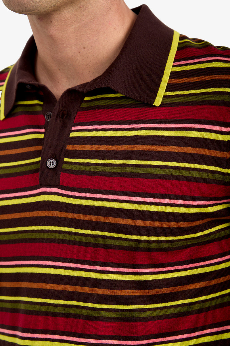 A close-up of a person wearing the Bayard Polo, which features a collared design and horizontal stripes in shades of red, yellow, pink, and brown. Only the lower face, neck, and upper torso are visible.