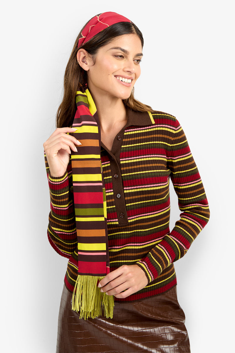 A woman with long brown hair smiles and looks to the side as she poses against a plain white background, wearing a brown skirt and a colorful striped sweater paired with the SERA FRINGE SCARF.