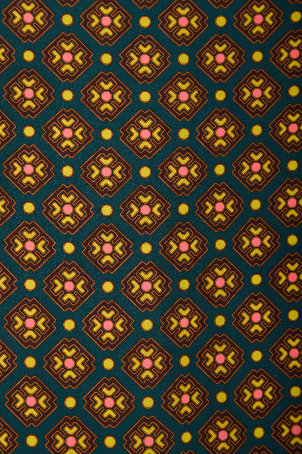 The FOULARD FRINGE SCARF features a repeating geometric pattern of diamond shapes with pink and yellow star-like centers and yellow dots, all set against a dark green background. This scarf showcases a retro, symmetrical style.