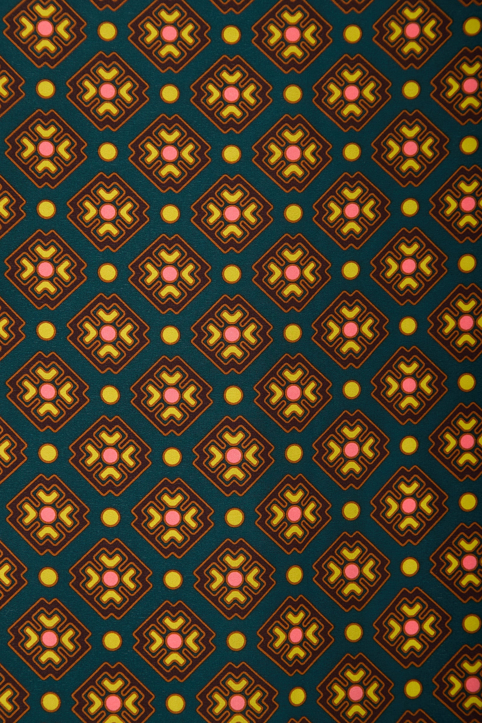 The FOULARD FRINGE SCARF features a repeating geometric pattern of diamond shapes with pink and yellow star-like centers and yellow dots, all set against a dark green background. This scarf showcases a retro, symmetrical style.