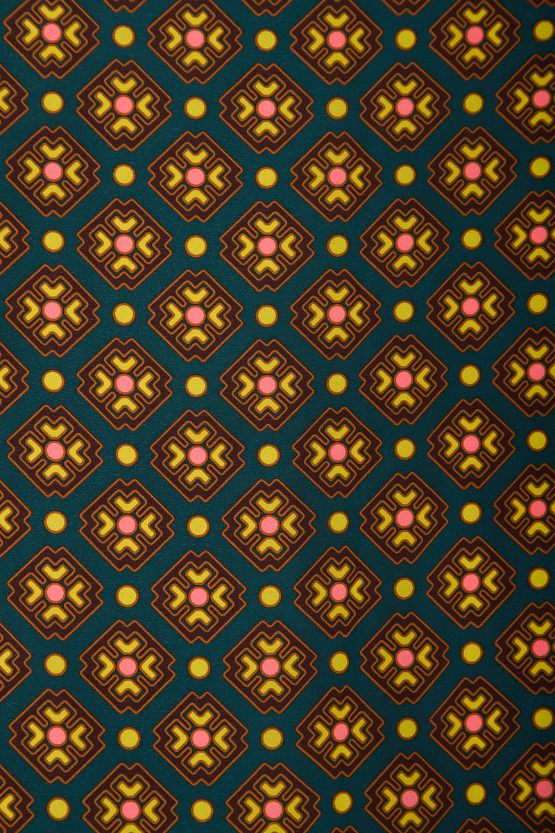 The FOULARD FRINGE SCARF features a repeating geometric pattern of diamond shapes with pink and yellow star-like centers and yellow dots, all set against a dark green background. This scarf showcases a retro, symmetrical style.