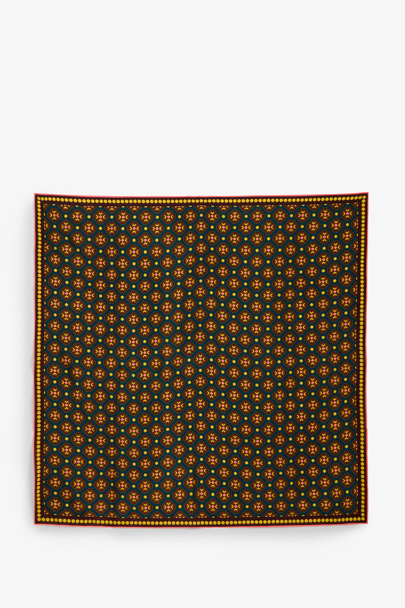 The FOULARD FRINGE SCARF features a symmetrical, evenly spaced repeating floral pattern in yellow and orange on a dark background, finished with a thin red and yellow border along the edge.