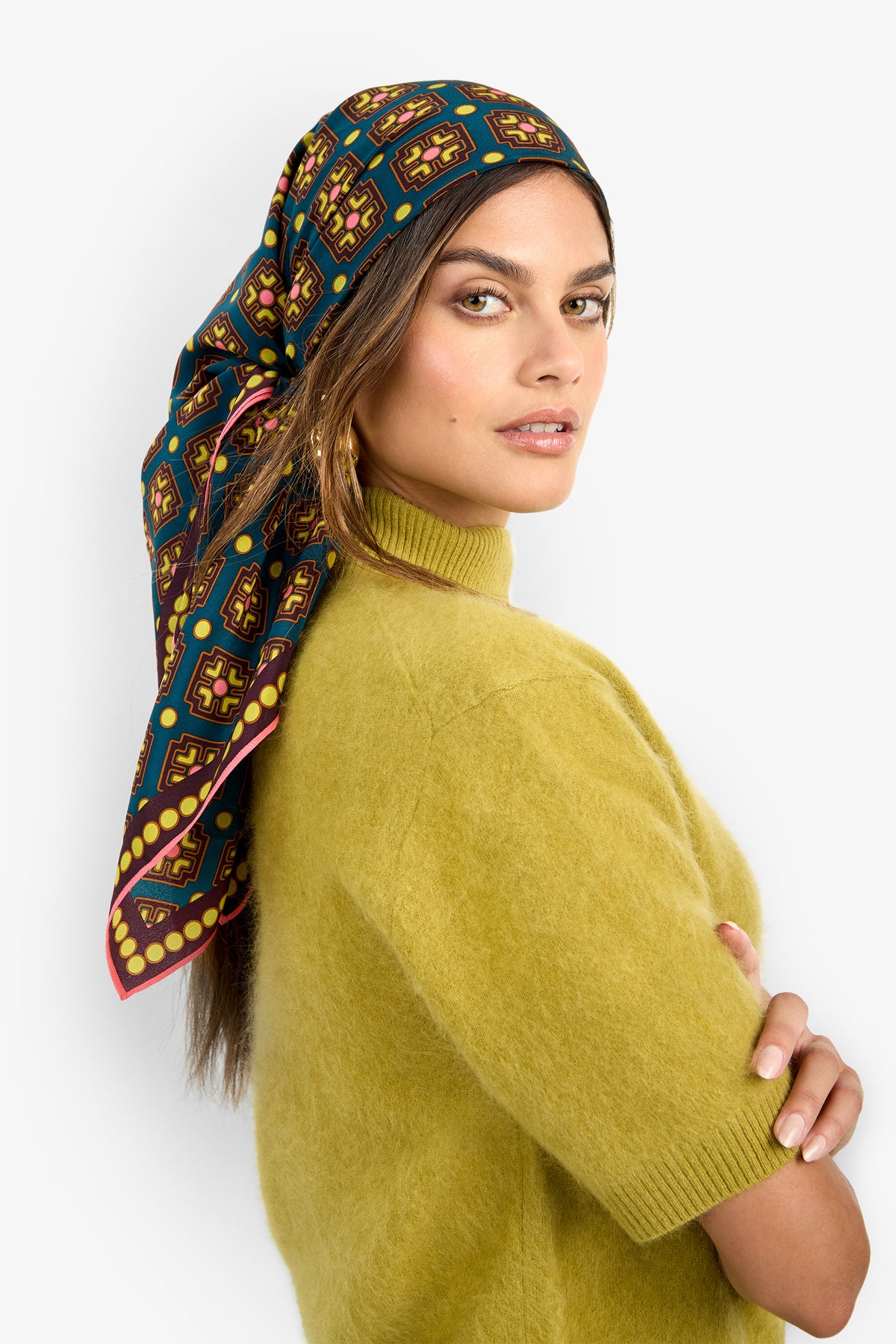 A woman stands sideways with her arms crossed against a plain light background, looking at the camera. She wears a yellow-green short-sleeve sweater and has the FOULARD FRINGE SCARF—featuring colorful geometric designs—styled over her hair.