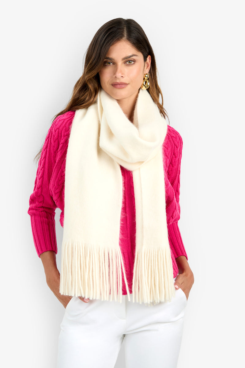 A person wearing a long, cream-colored Carducci Oversized Scarf with fringe ends over a bright pink cable-knit sweater and white trousers.
