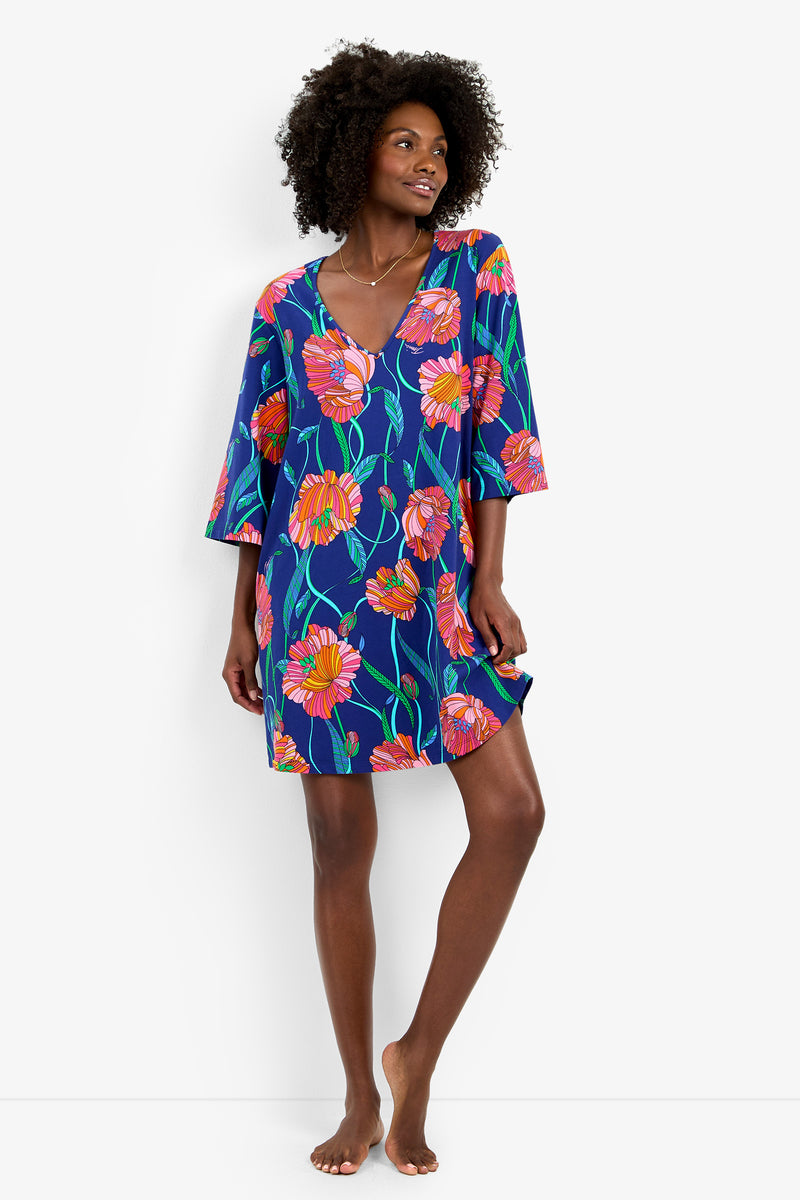 A woman stands barefoot against a plain white background, smiling and looking to the side while wearing the Linear Poppy Sleepshirt—a blue floral dress adorned with pink and orange flowers.