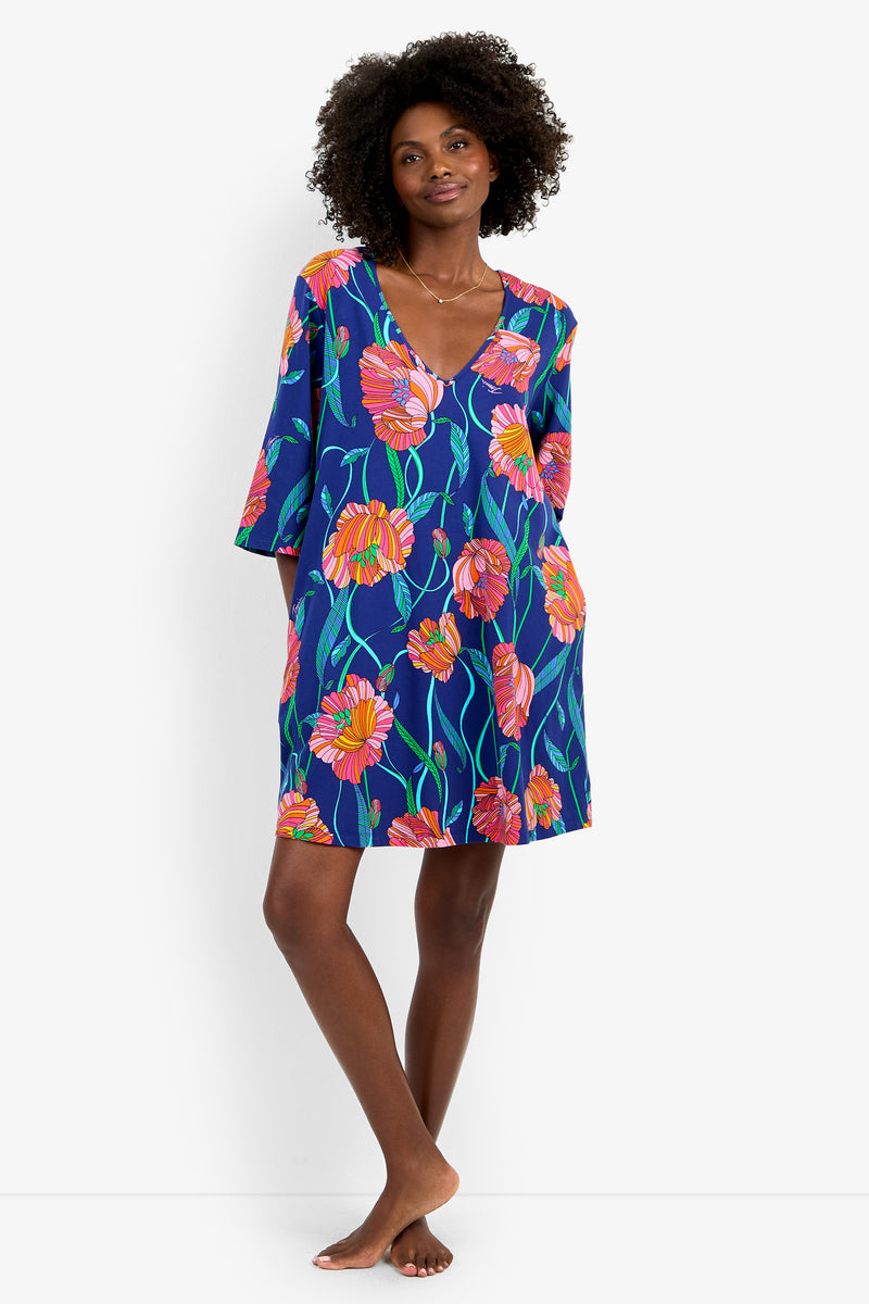 A woman with curly hair stands barefoot, smiling as she wears the Linear Poppy Sleepshirt—a blue dress featuring bright pink and orange poppy floral prints, a V-neckline, three-quarter sleeves, and convenient side pockets.