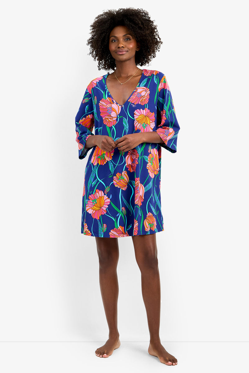 A woman stands barefoot against a plain white background, wearing the Linear Poppy Sleepshirt—a loose, knee-length blue dress featuring vibrant orange and pink floral patterns.