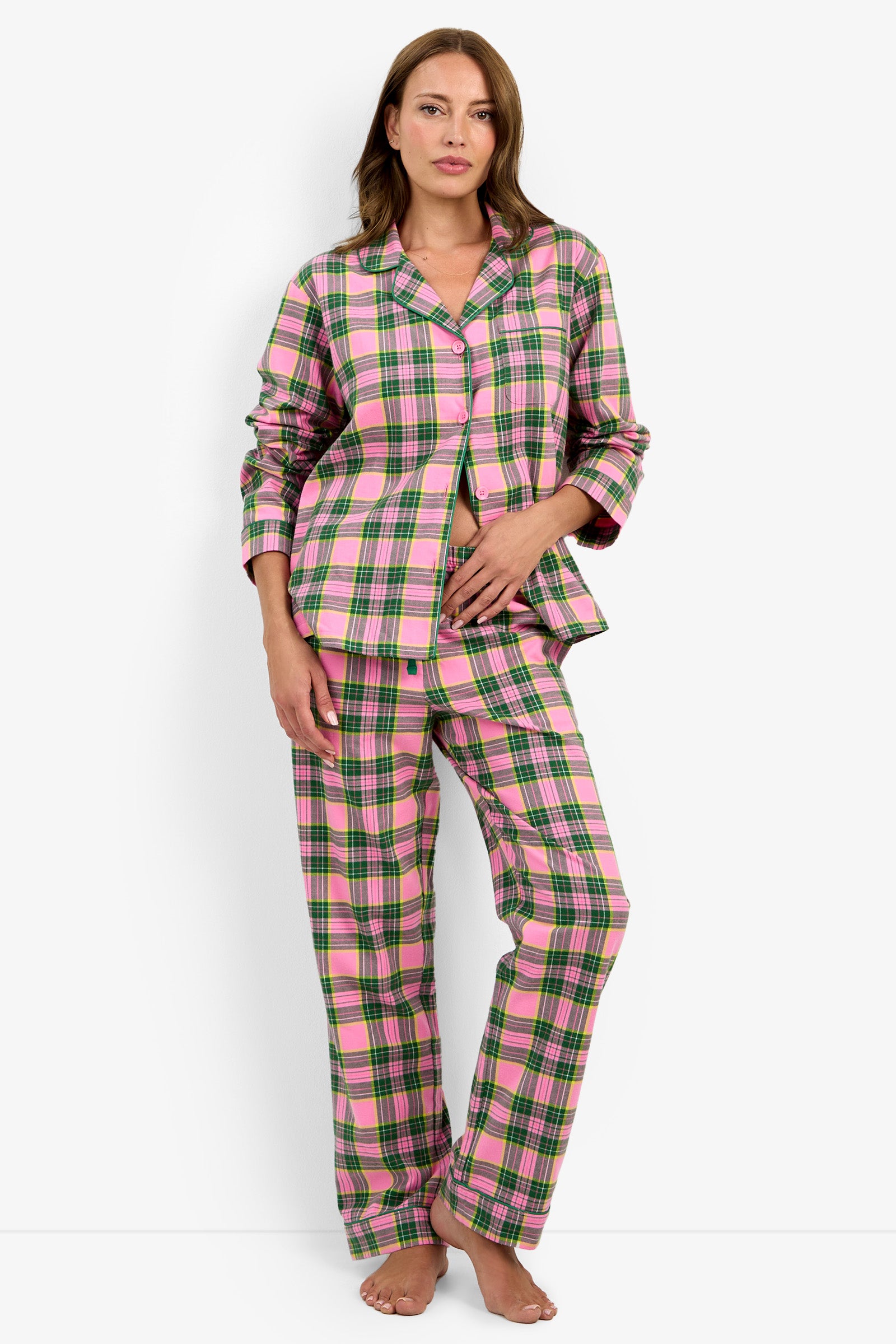 A woman stands barefoot against a plain white background, wearing the Tartan Long Sleeve PJ Set—a pink, green, and white plaid pajama set featuring long sleeves and long pants. One hand rests on her stomach while the other hangs at her side.