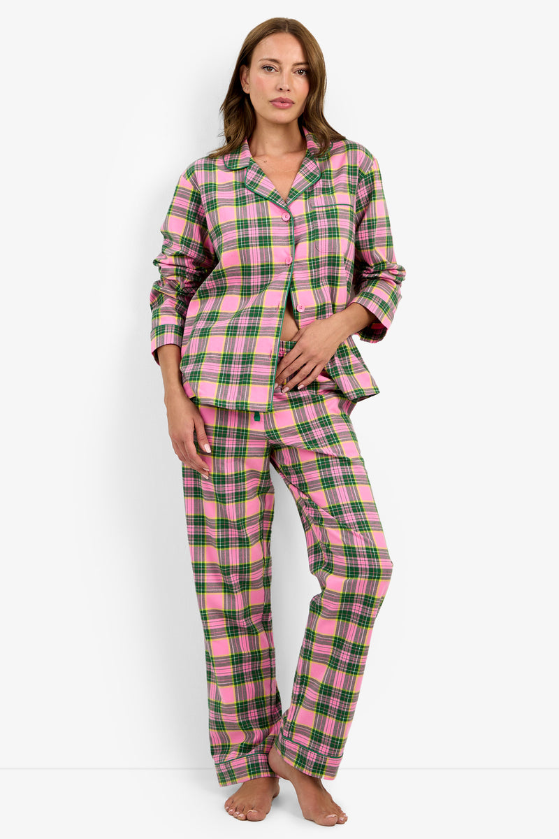 A woman stands barefoot against a plain white background, wearing the Tartan Long Sleeve PJ Set—a pink, green, and white plaid pajama set featuring long sleeves and long pants. One hand rests on her stomach while the other hangs at her side.