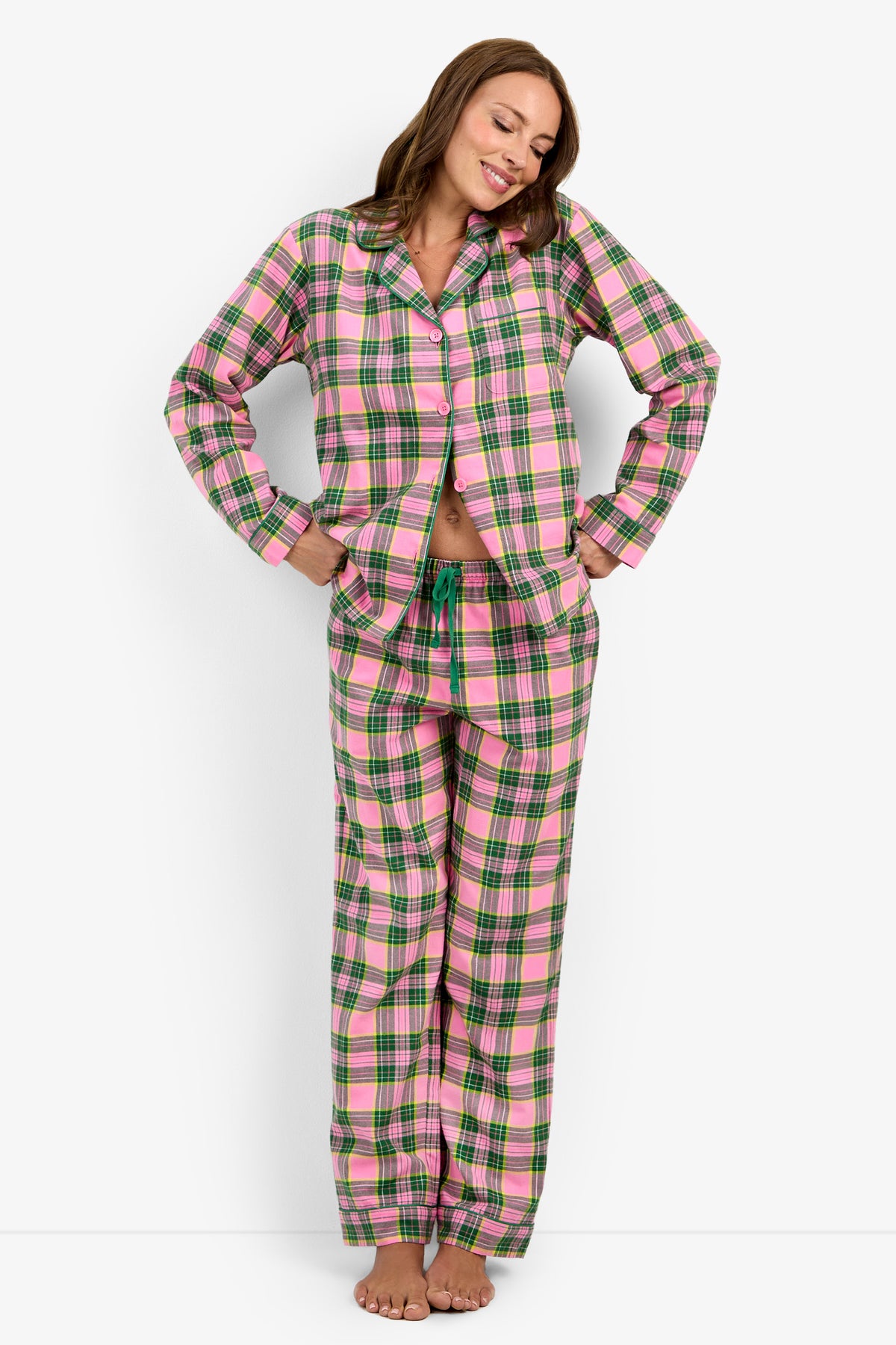 A woman stands barefoot against a white background, smiling as she wears the Tartan Long Sleeve PJ Set—featuring a pink, green, and white plaid pattern with long sleeves and matching pants—her hands resting on her hips.