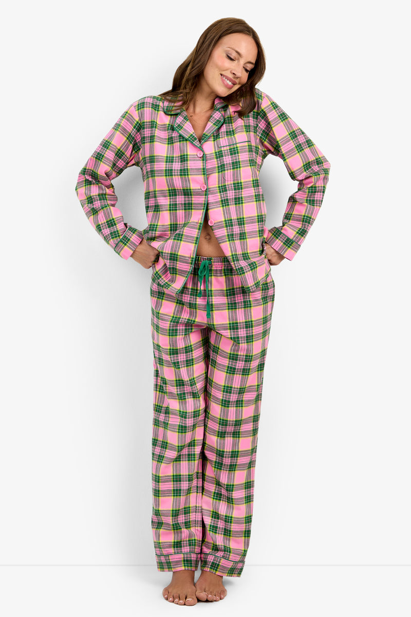 A woman stands barefoot against a white background, smiling as she wears the Tartan Long Sleeve PJ Set—featuring a pink, green, and white plaid pattern with long sleeves and matching pants—her hands resting on her hips.