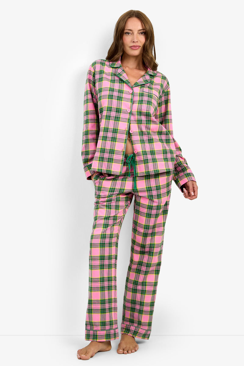A woman models the Tartan Long Sleeve PJ Set—loose-fitting pajamas in a pink, green, and white plaid—standing barefoot against a white background with one hand in her pocket and a relaxed expression.