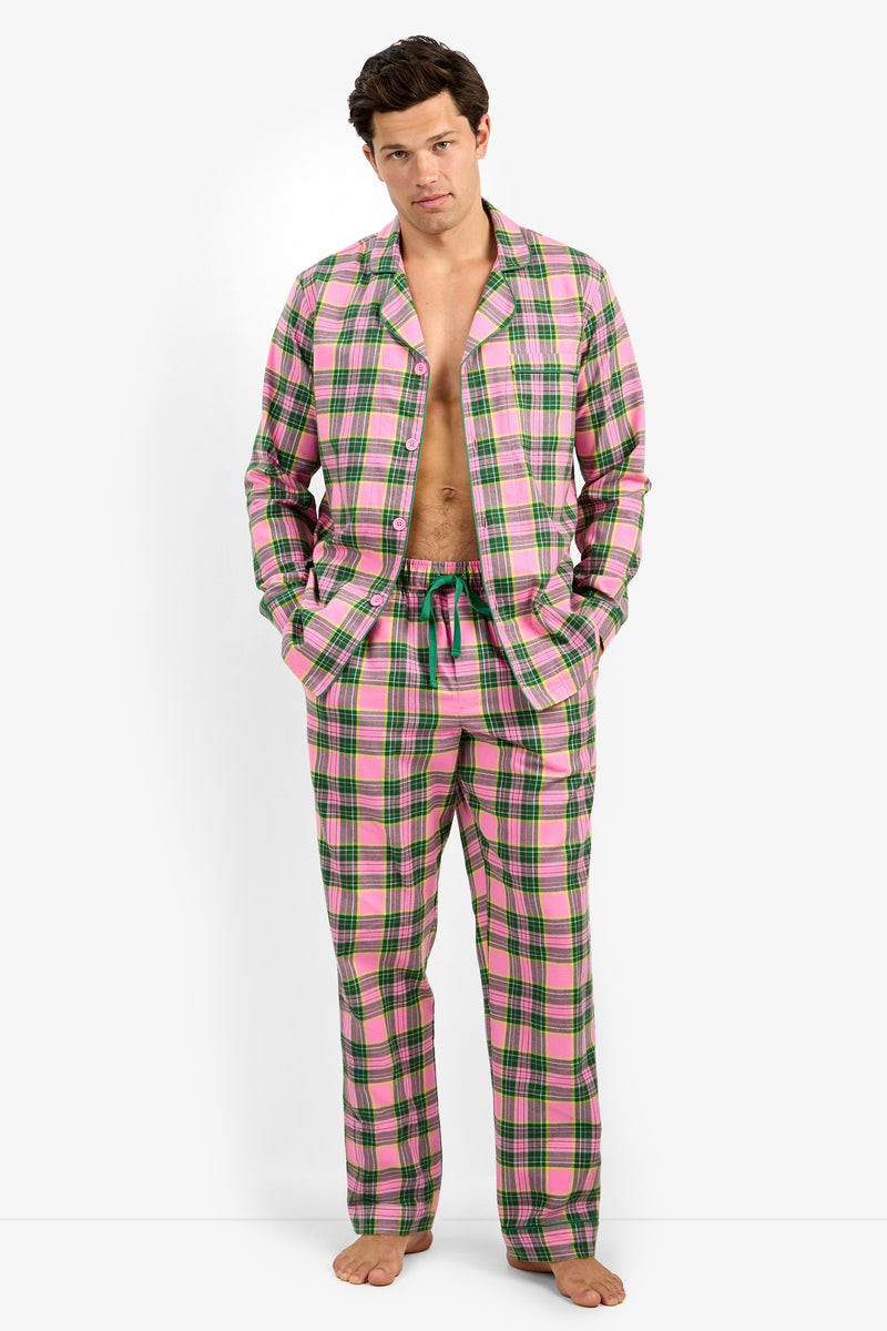 A man modeling a pink and green plaid pajama set, consisting of a long-sleeve open shirt and matching pants, against a plain white studio background.