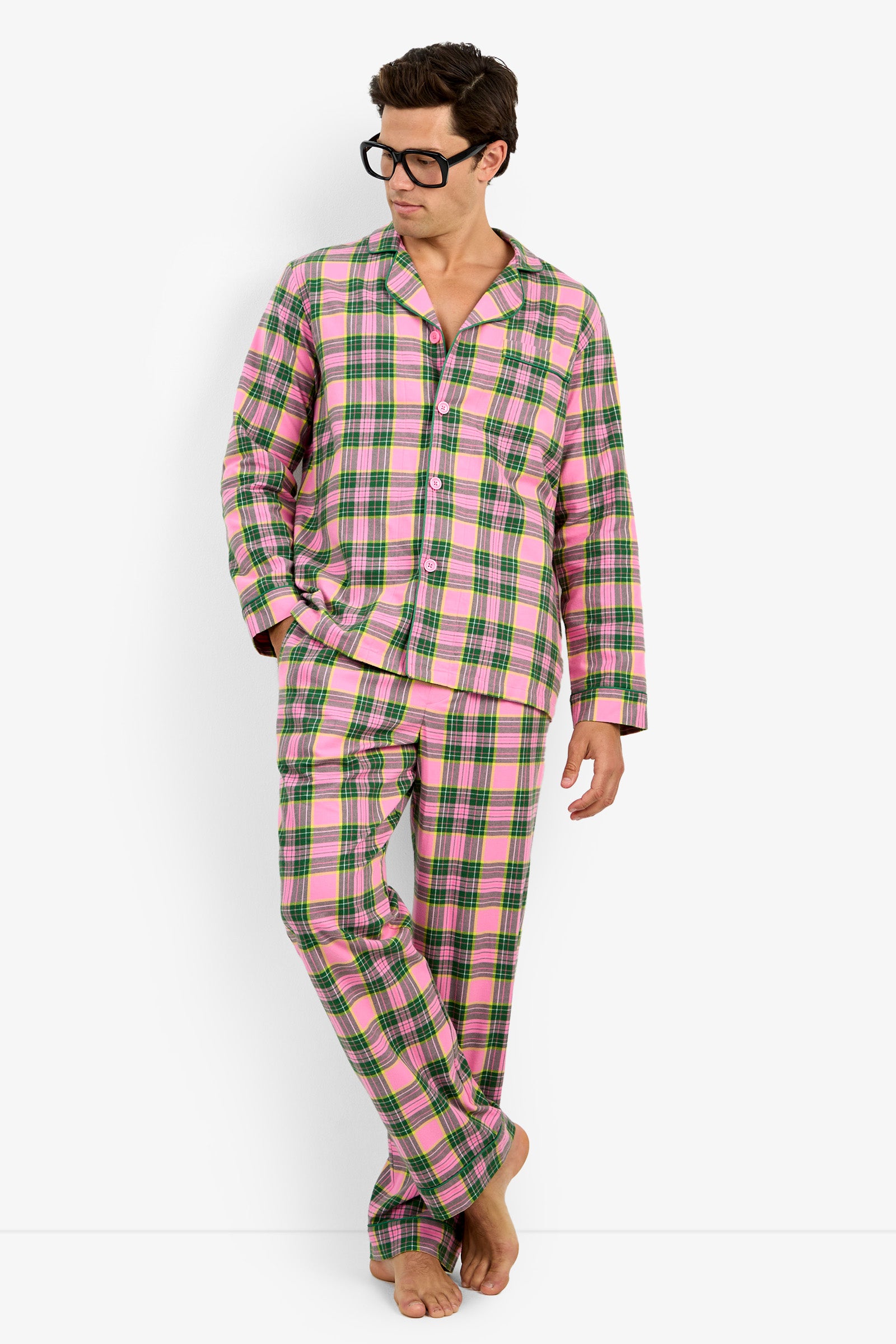 A man wearing a pink, green, and white plaid pajama set with black-rimmed glasses, standing against a plain white studio background.