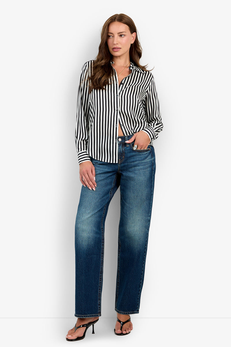 A woman with long brown hair stands in front of a white background, wearing the Rouge Top—a black and white vertical striped button-down shirt—paired with blue jeans and black heeled sandals. She poses in a relaxed manner.