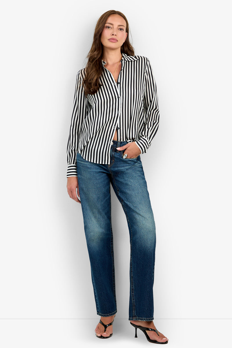 A woman stands against a white background wearing the Rouge Top—a black and white vertical striped button-up shirt—paired with blue jeans and black heeled sandals. She has long brown hair, a neutral expression, and one hand in her pocket.
