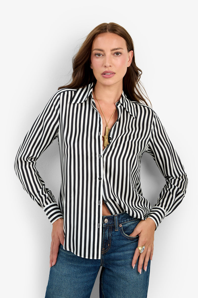 A woman poses against a plain light background, wearing the Rouge Top—a black and white vertically striped button-up shirt—paired with blue jeans. She has her hand in her pocket and loose brown hair.