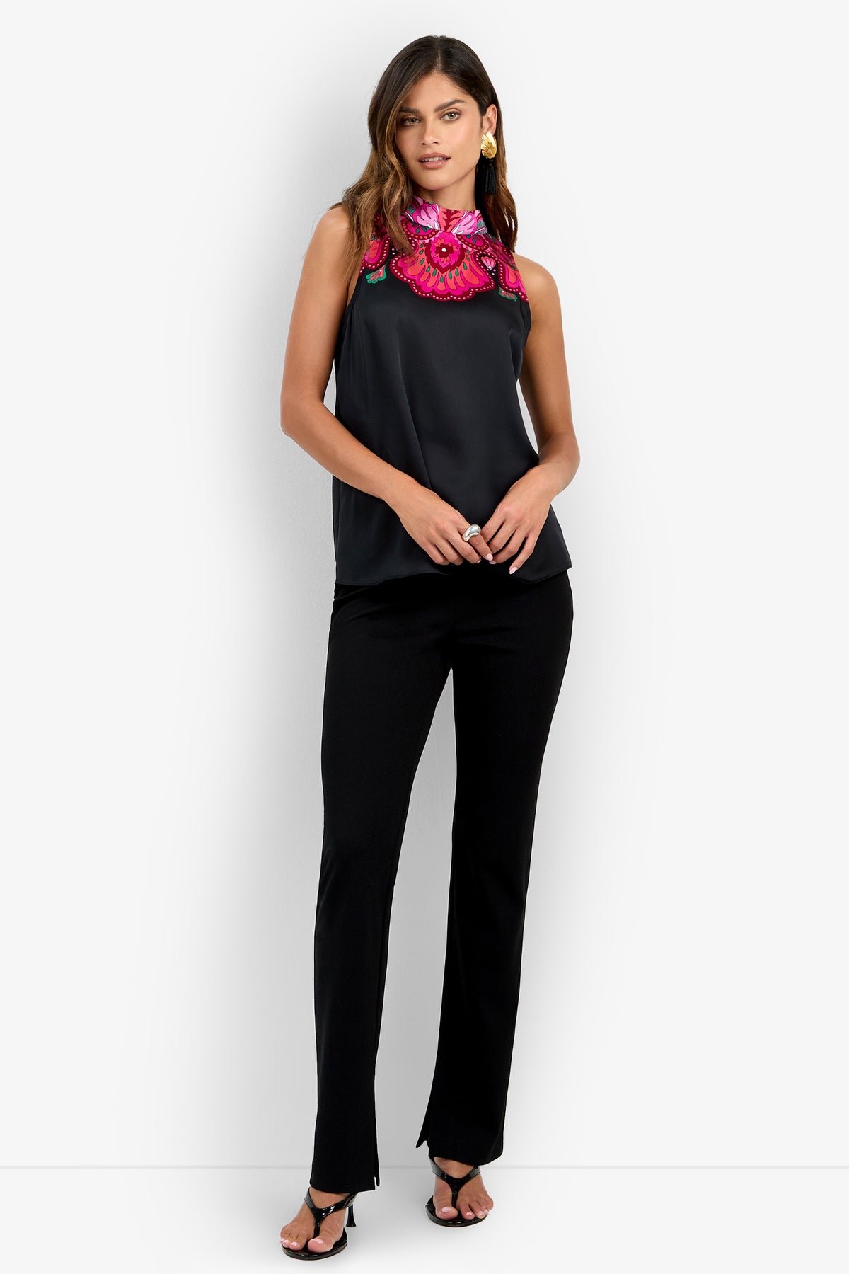 A woman stands against a plain white background wearing the Loveage Top in Sautoir, featuring a sleeveless black design with a large, bright pink floral embroidered collar. She pairs it with black pants, black heels, and gold earrings.