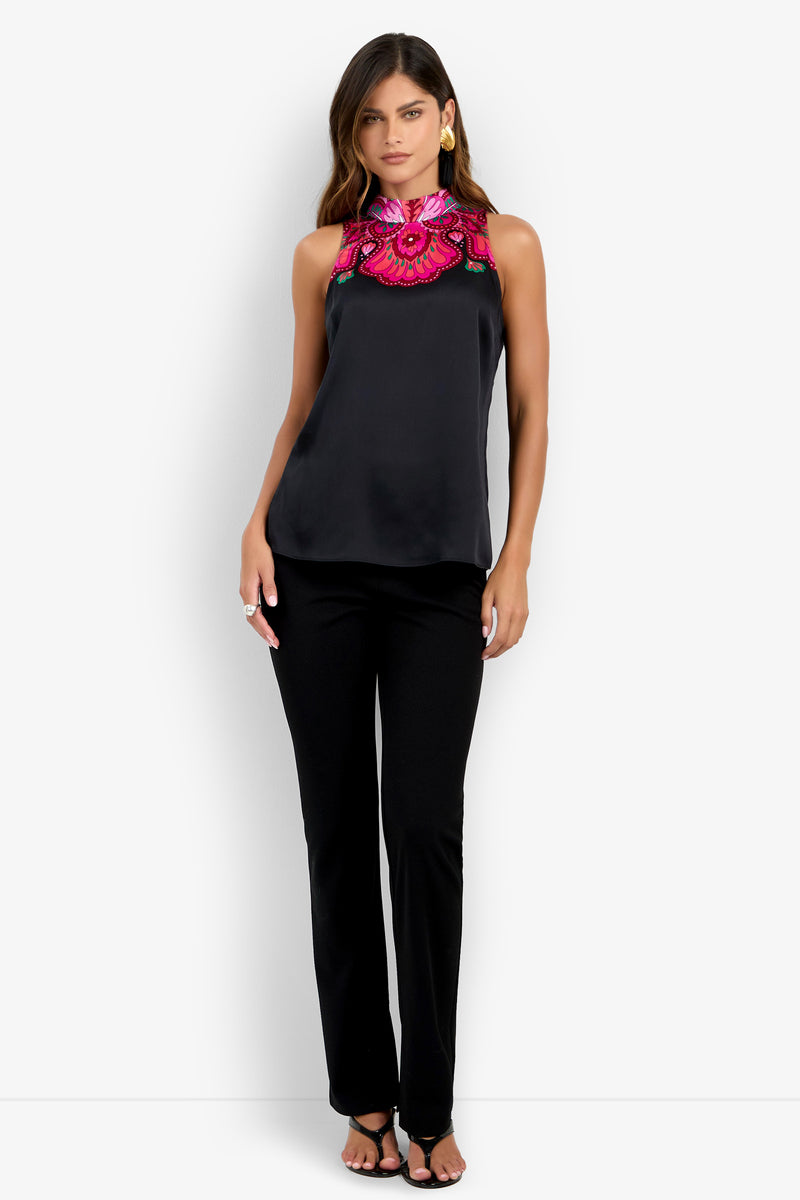 A woman is standing and facing forward with one hand on her hip, wearing the Loveage Top—a black sleeveless top featuring bright pink floral embroidery on the collar—paired with black pants.